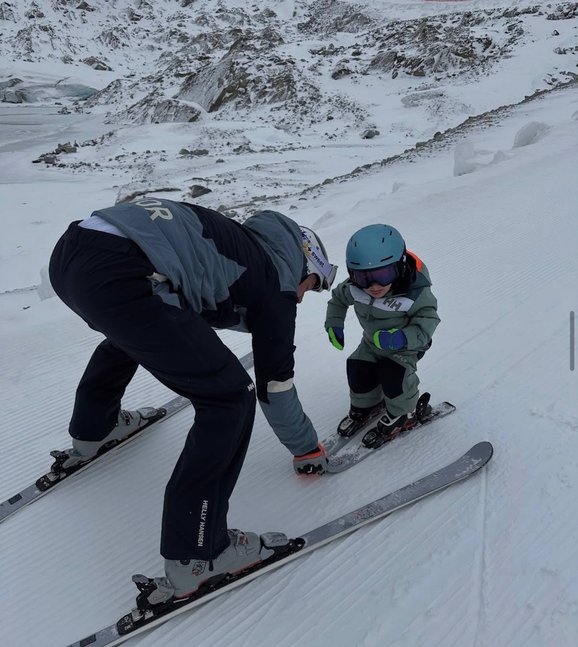 En voksen person hjelper et barn med å feste skiene i et snødekket fjellandskap. (Bildebeskrivelsen er laget av en KI-tjeneste)