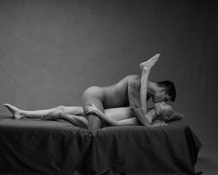 En naken dame ligger på ryggen i senga med venstre ben opp langs overkroppen til en naken mann som ligger oppå henne mens de kysser