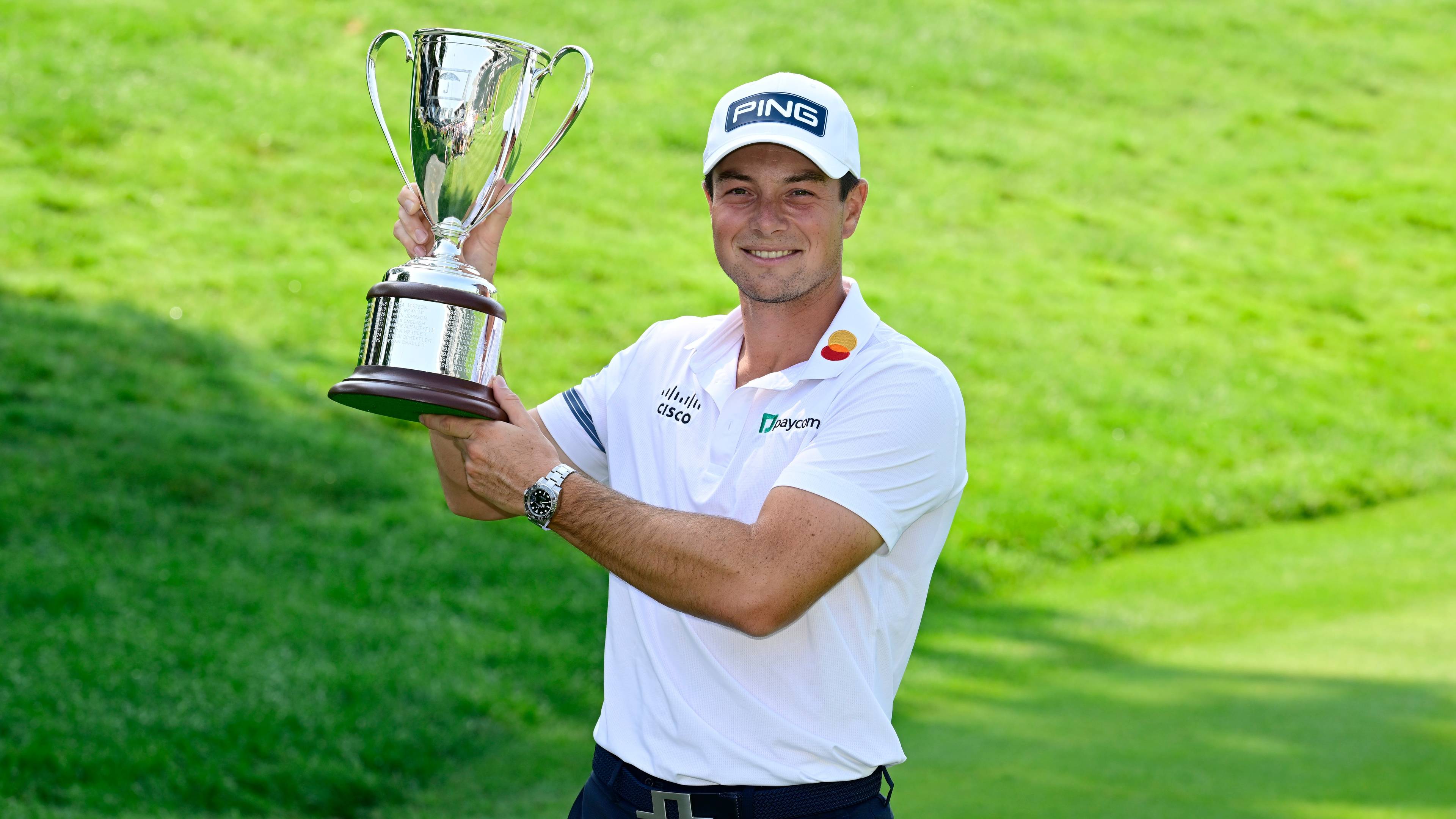 Viktor Hovland smiler og holder en golfpokal på en gressbane. (Bildebeskrivelsen er laget av en KI-tjeneste)