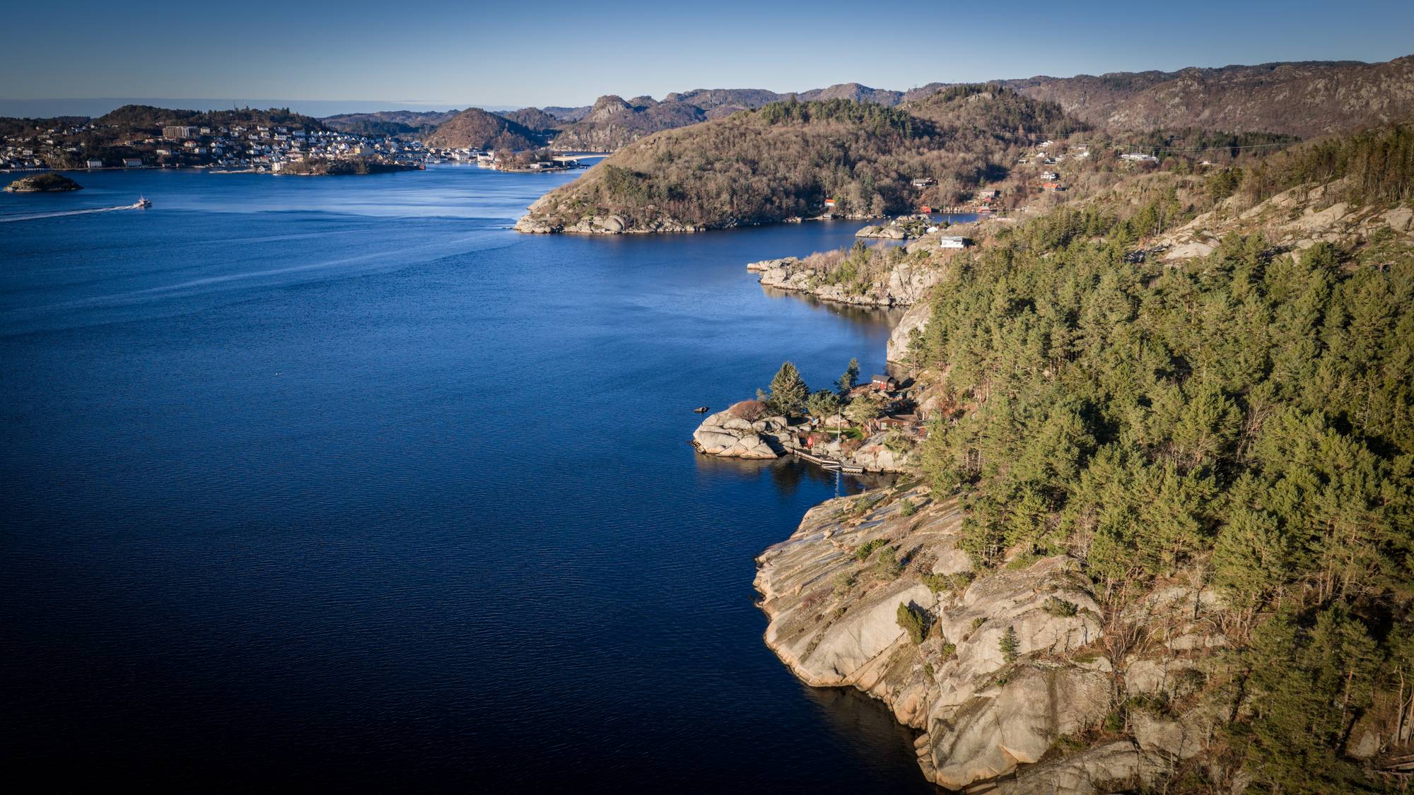 Bildet viser en kystlinje med klipper og furuskog langs en rolig sjø. Flere boligområder er synlige på bakgrunnen av båter som ligger til havn. Lyset er klart, og det er rolige vannforhold. Omgivelsene gir en naturpreget atmosfære med fjell i det fjerne. (Bildebeskrivelsen er laget av en KI-tjeneste)