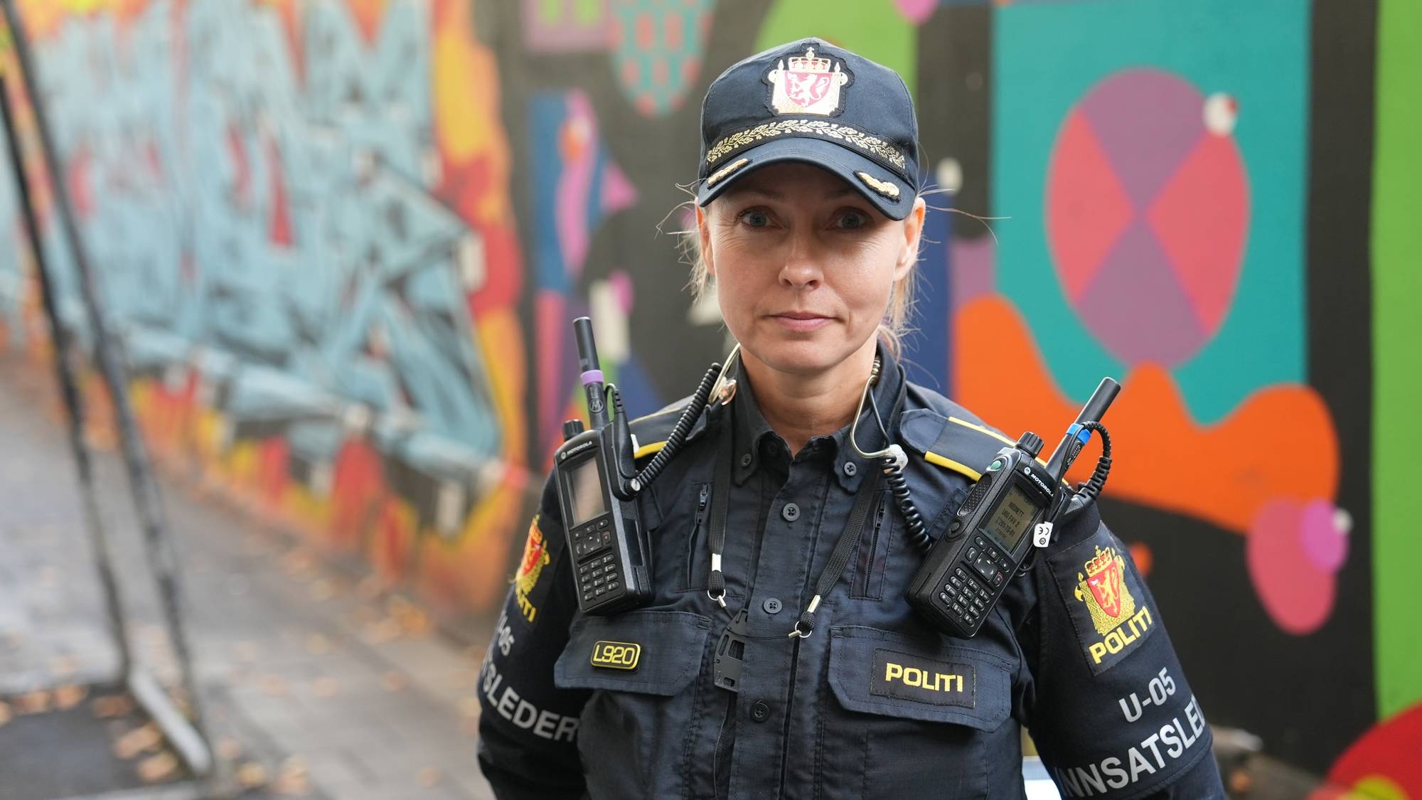 Kvinnelig politibetjent Ine Jahren i uniform med samband foran en graffiti-vegg. (Bildebeskrivelsen er laget av en KI-tjeneste)