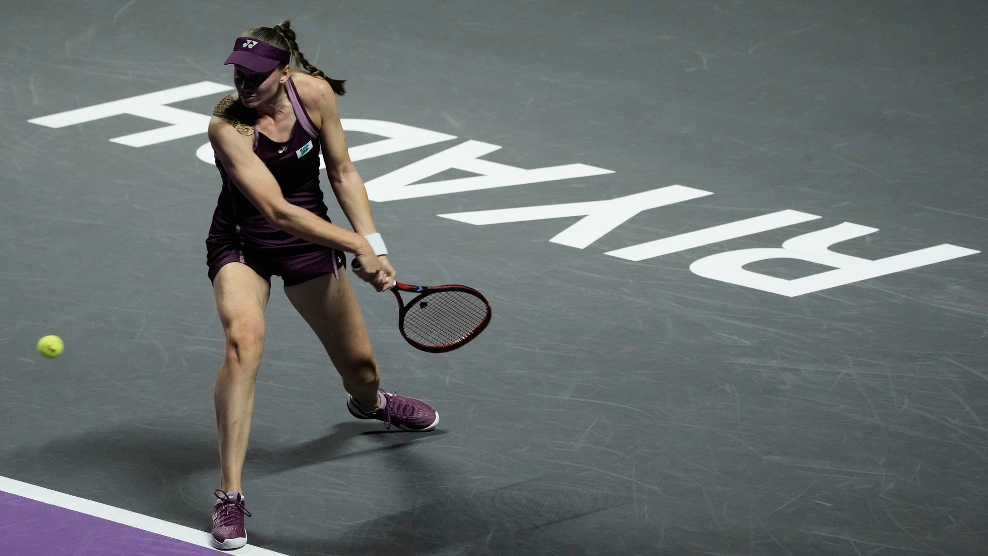 En kvinnelig tennisspiller med lilla drakt slår til ballen på en mørk bane. Bak henne er store hvite bokstaver synlige. Hun bruker en rød racket. Ballen er i luften til venstre for henne. (Bildebeskrivelsen er laget av en KI-tjeneste)