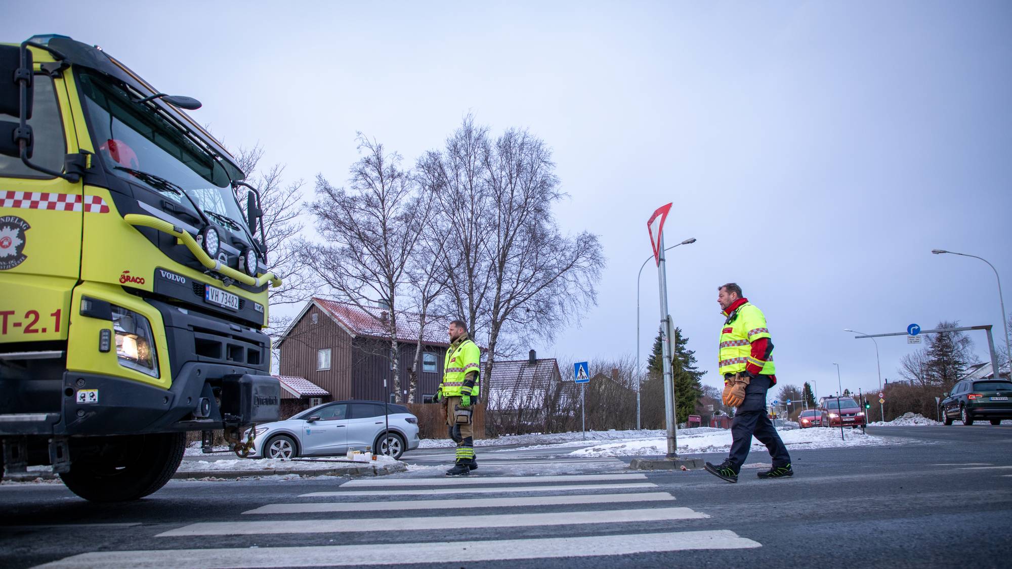To brannmenn i refleksjakker står ved en fotgjengerovergang. En brannbil er parkert foran dem. Bakgrunnen viser hus og snødekte veier. Det er overskyet vær.