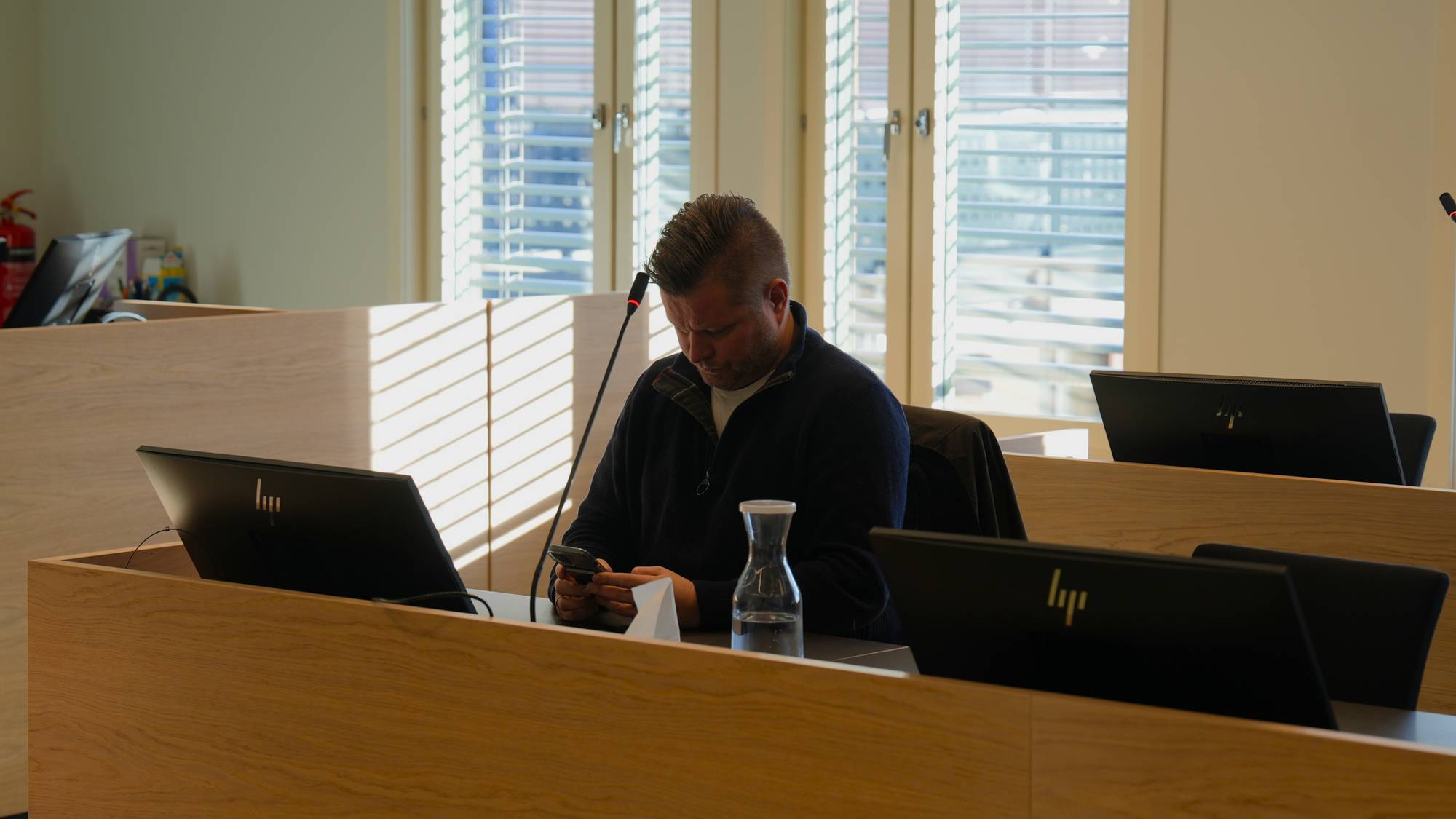 Mann sitter ved en pult i en rettssal og ser på en mobiltelefon. (Bildebeskrivelsen er laget av en KI-tjeneste)