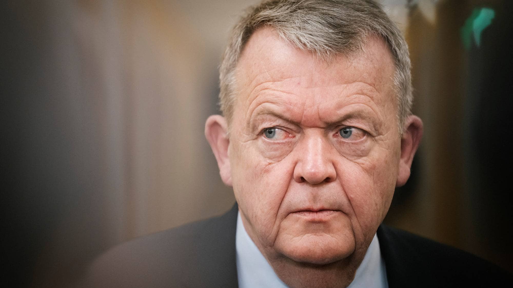 Dansk politiker Lars Løkke Rasmussen ser til siden. Han er kledd i mørk dress og lys skjorte. Bildet er et portrett av mannen. Omgivelsene er mørke og uskarpe. (Bildebeskrivelsen er laget av en KI-tjeneste)