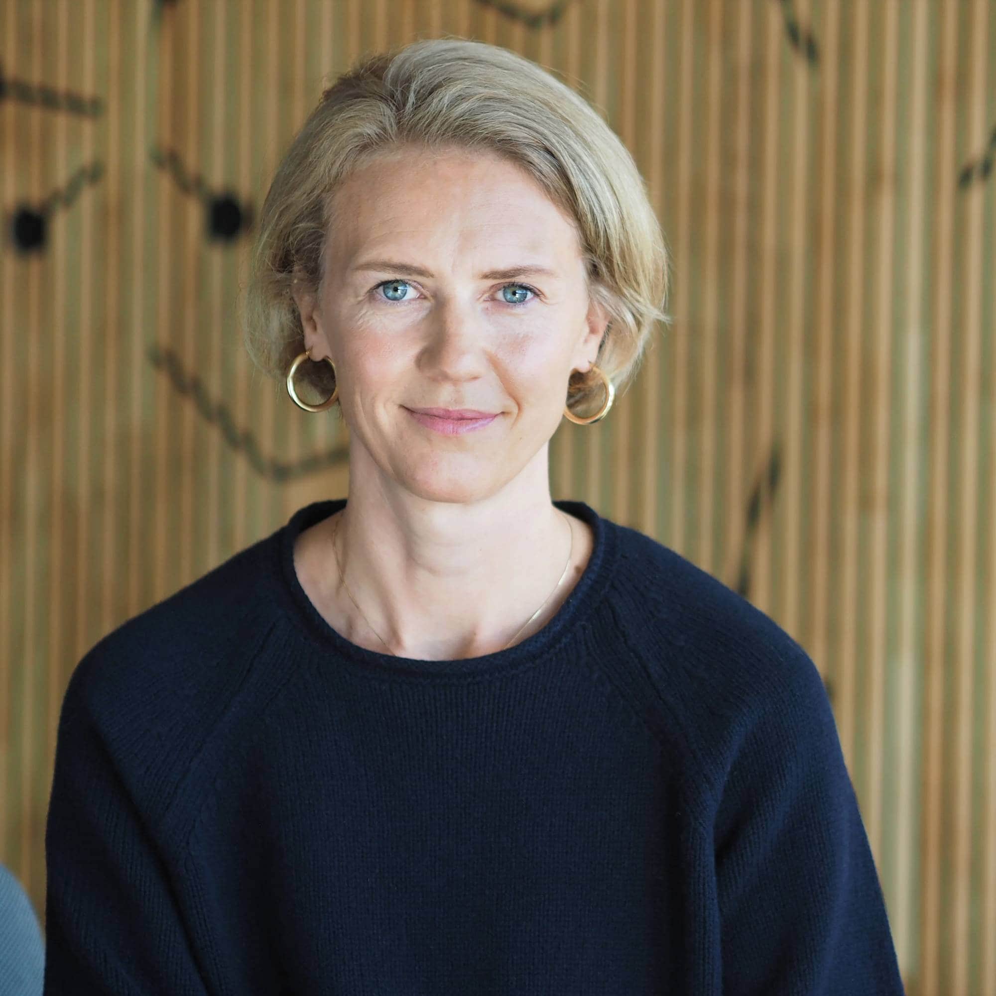 Overlege Ingrid Aas smiler forsiktig til kamera. Hun har kort, lyst hår, blå øyne og lys hud. Hun har på seg en svart genser.