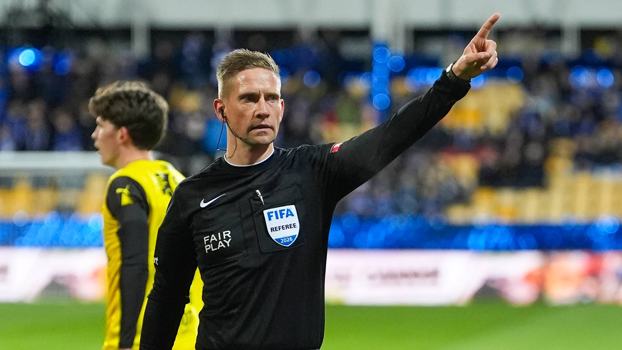 En fotballdommer peker med armen på en bane under en kamp. En spiller i gul drakt står i bakgrunnen. (Bildebeskrivelsen er laget av en KI-tjeneste)