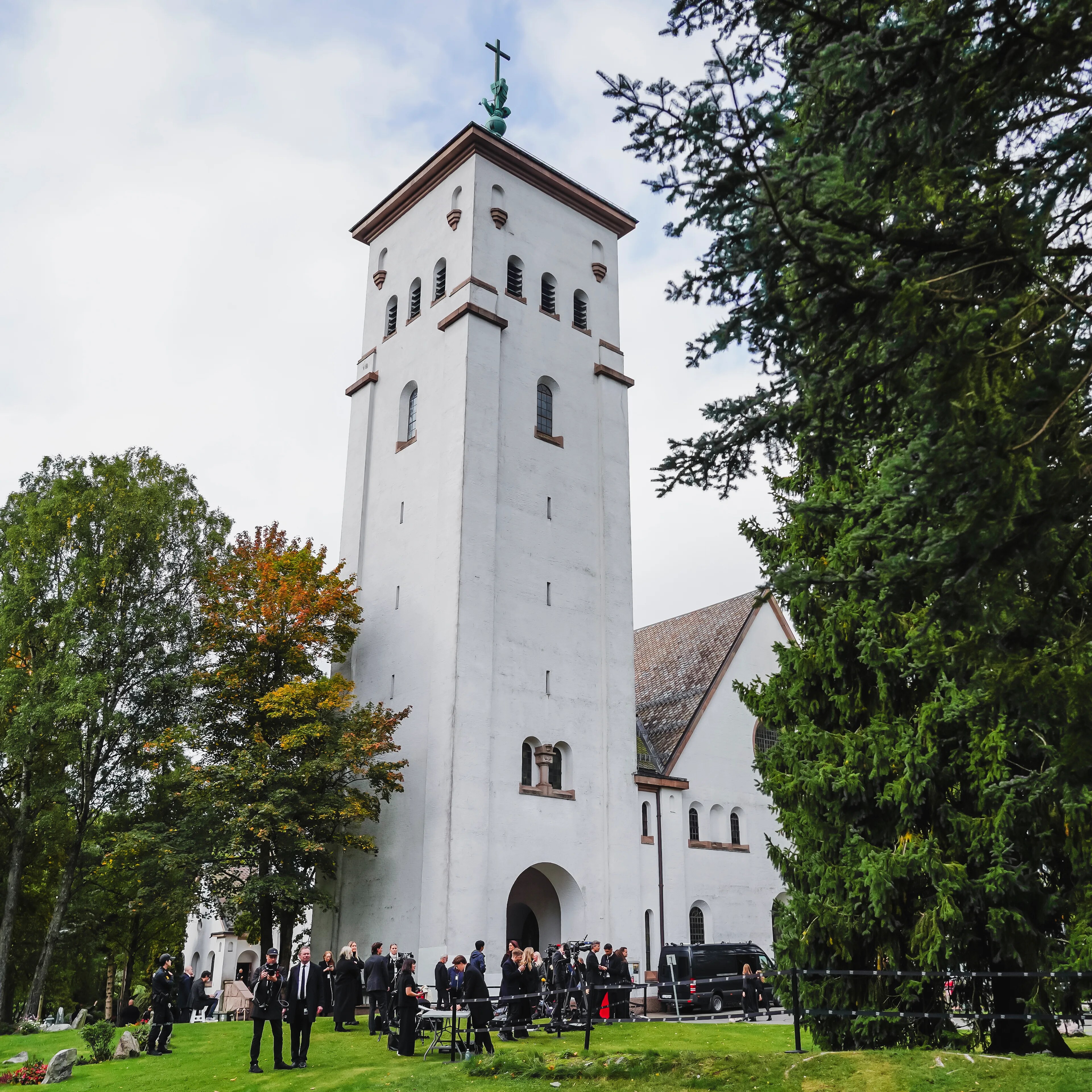 en hvit kirke fortografert på avstand, foran kirka står en folkemengde kledd i sort