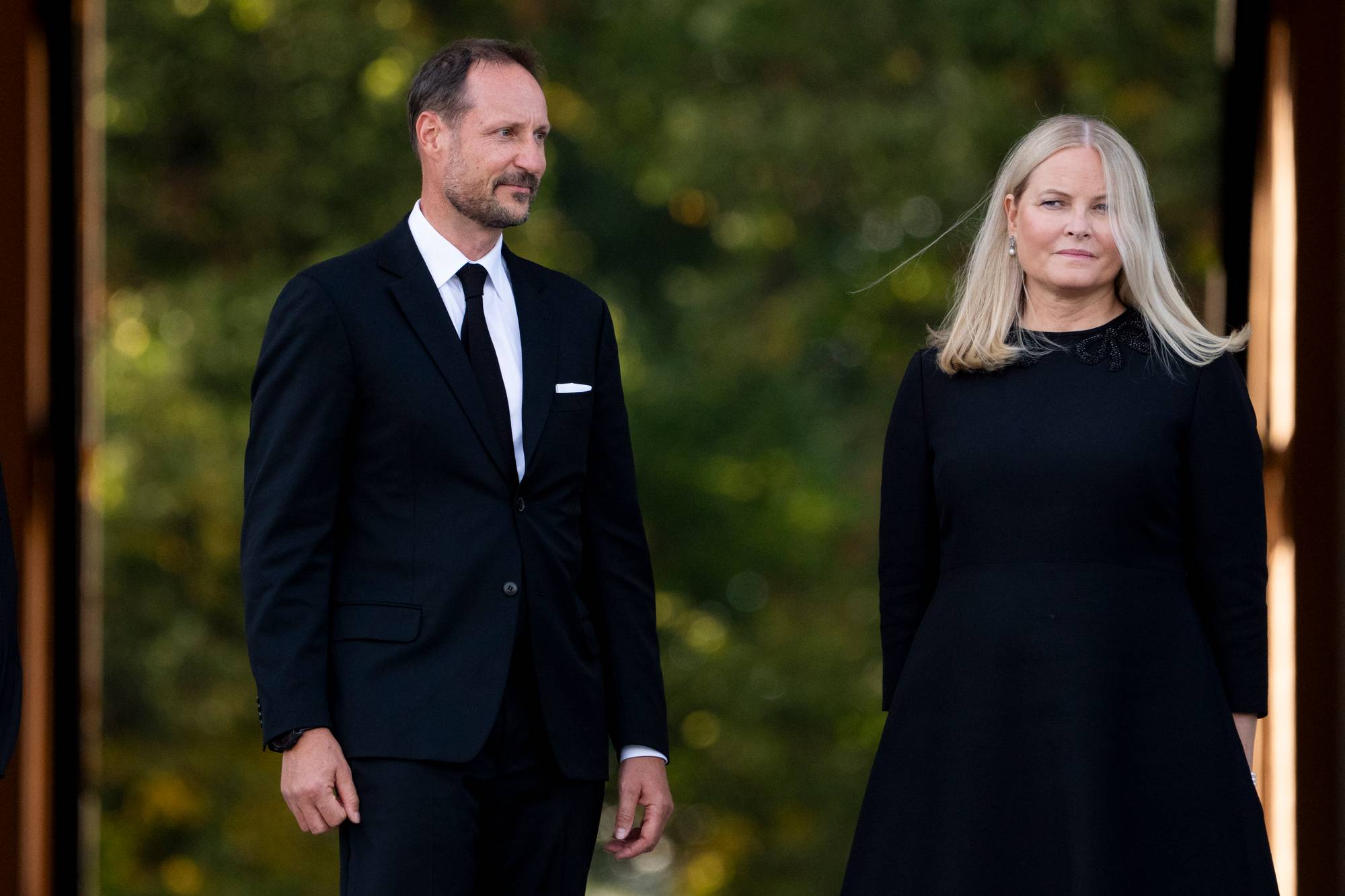 Kong Haakon og dronning Mette-Marit med i svarte klær med alvorlige ansiktsuttrykk.