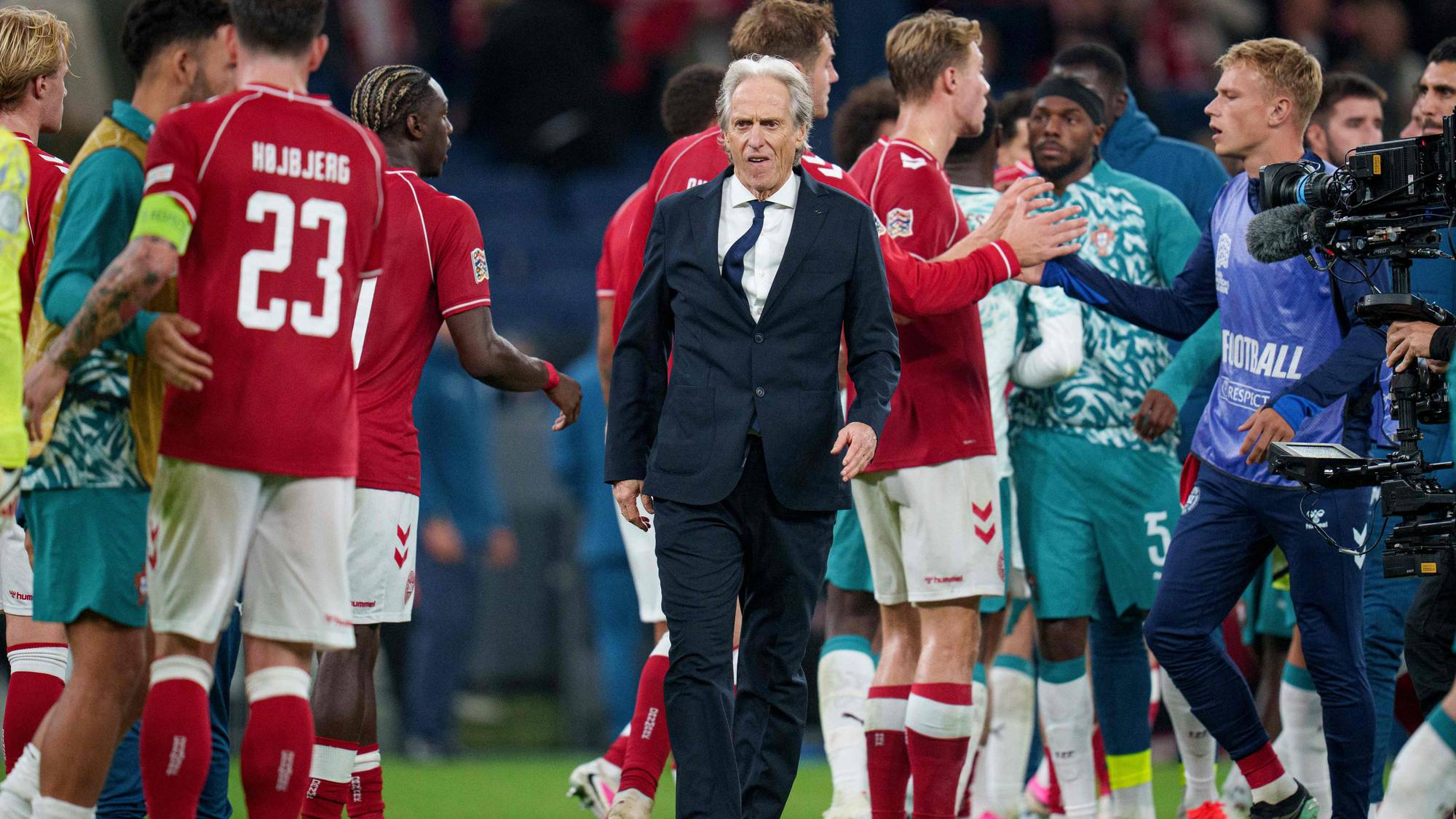 Fotballtrener i dress går over banen omgitt av spillere i røde og grønne drakter etter kamp. (Bildebeskrivelsen er laget av en KI-tjeneste)
