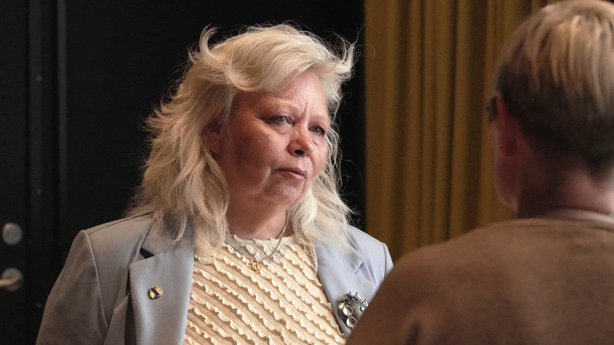 En kvinne med hvitt, bølgete hår og en lyseblå blazer ser til siden. Hun har en beige overdel med et mønster og et smykke rundt halsen. En person med brun genser er sett fra ryggen. (Bildebeskrivelsen er laget av en KI-tjeneste)
