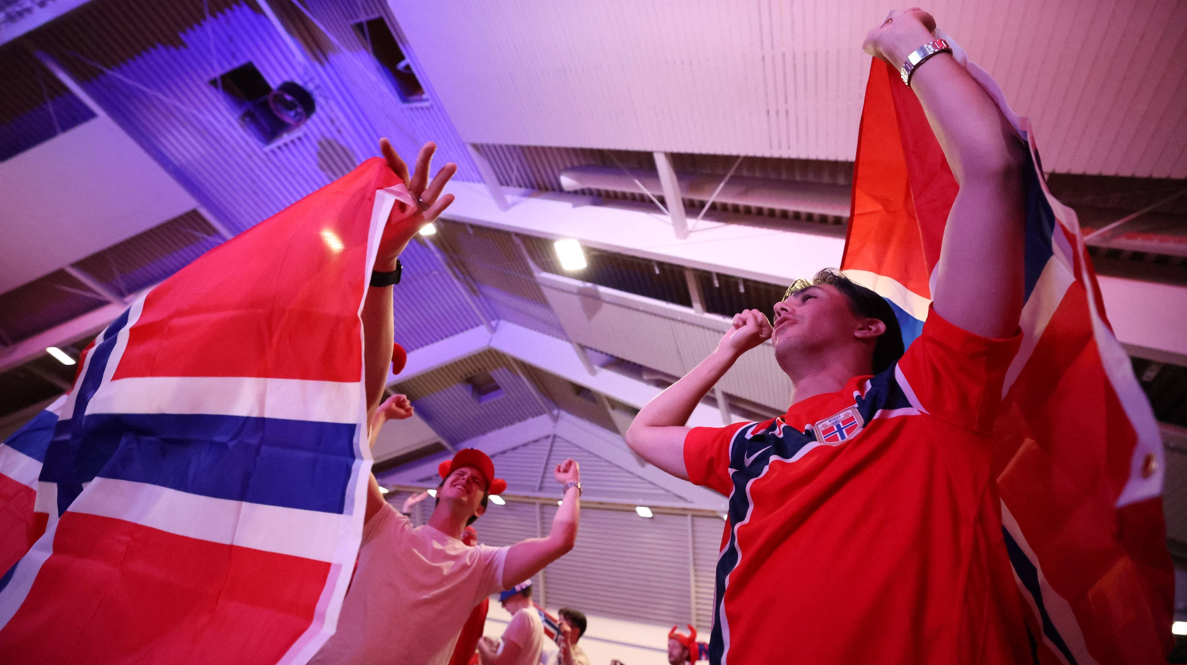 Folk i røde supportertrøyer holder norske flagg. En person til høyre holder et norsk flagg og blåser i en fløyte. Til venstre holder en annen person et norsk flagg. Bakgrunnen er et tak med lyssetting. (Bildebeskrivelsen er laget av en KI-tjeneste)