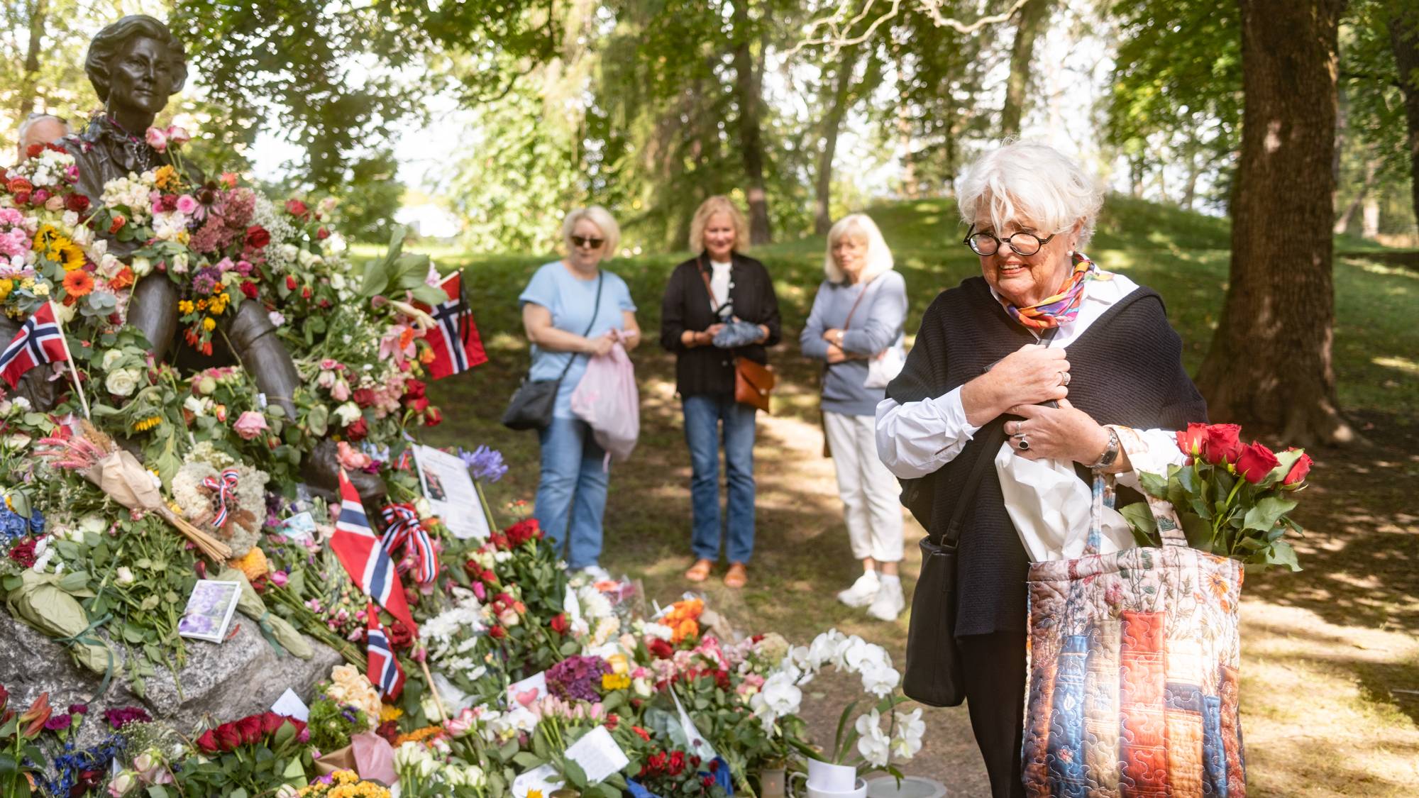 En dame ser rørt ut etter at hun akkurat har lagt ned en bukett blomster ved statuen av dronning Sonja. Det er et hav av blomster og lys, og mange mennesker som ser på blomstene.
