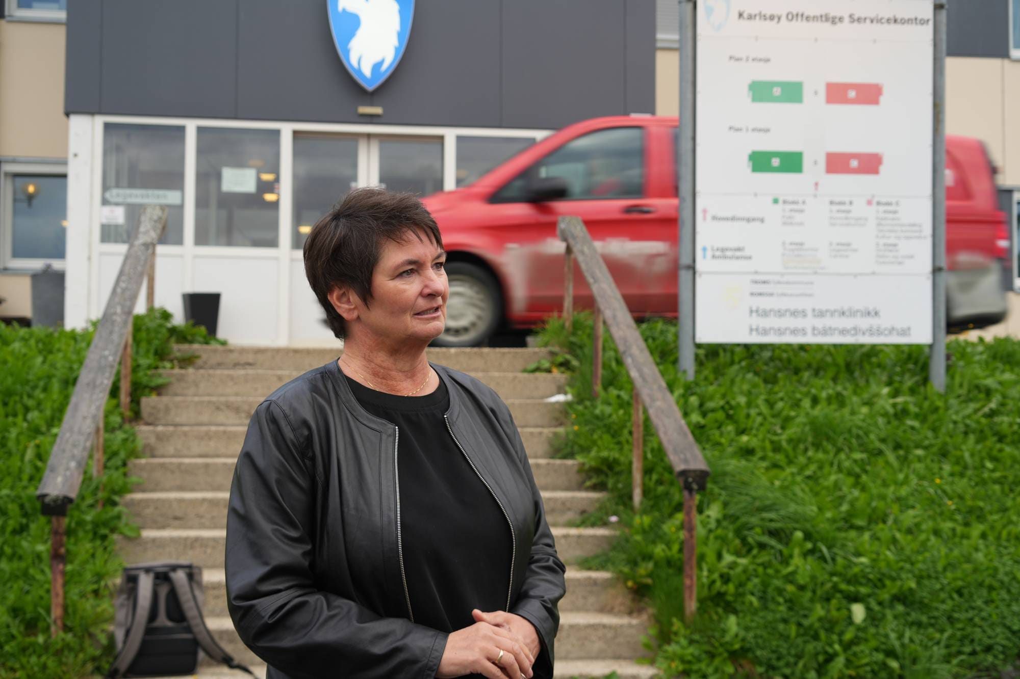 Kvinne står foran Karlsøy offentlige servicekontor. Bak henne sees en trapp og en rød bil. (Bildebeskrivelsen er laget av en KI-tjeneste)
