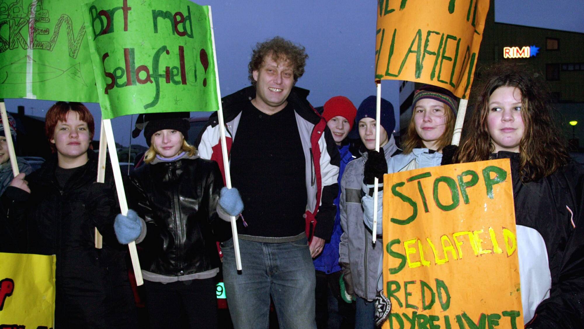 Demonstranter holder skilt med slagord som "Bort med Sellafield!" og "Stopp Sellafield".
En mann og flere ungdommer deltar i demonstrasjonen.
Bakgrunnen viser en bygning med et Rimi-skilt.
Bildet er tatt utendørs i skumringen. (Bildebeskrivelsen er laget av en KI-tjeneste)