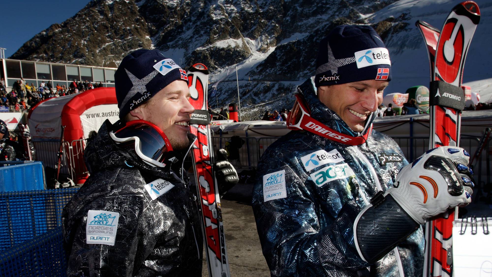 To skiløpere står side om side med ski på ryggen. Begge har på seg luer med norsk flagg og sponsorenes logoer. I bakgrunnen ses en bygning og snødekte fjell. (Bildebeskrivelsen er laget av en KI-tjeneste)
