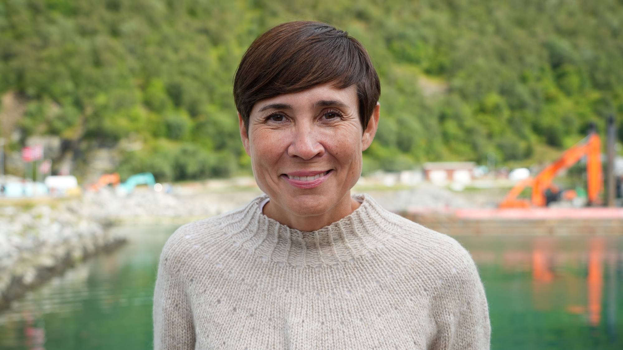 En dame med kort brunt hår ser smilende inn i kameraet. Hun har på en beige strikkegenser. Bildet er tatt ved sjøen.