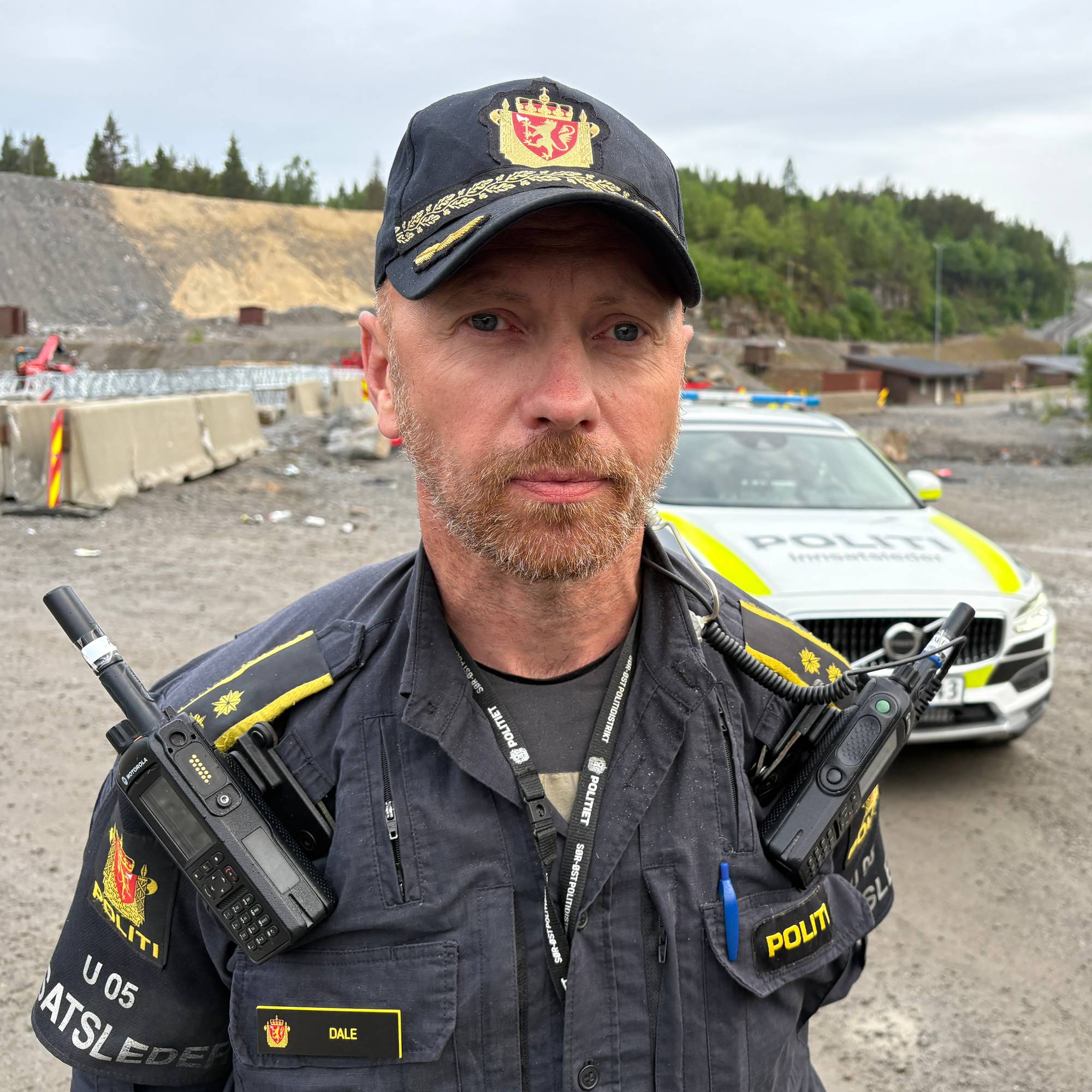 En politimann står med to sambandsradioer festet til uniformen. En politibil er synlig i bakgrunnen. 