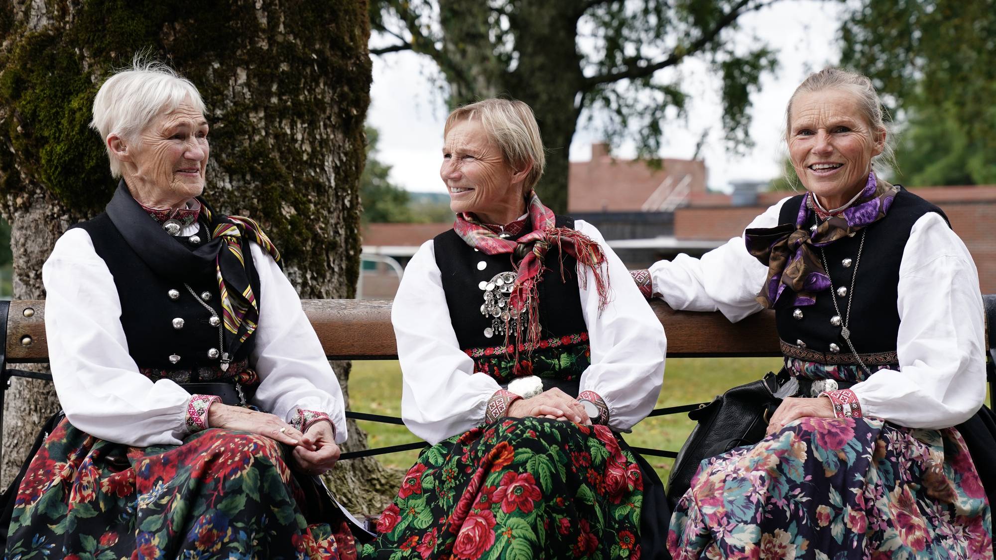 Tre voksne kvinner har på seg bunad og sitter på en benk utenfor en kirke. 
