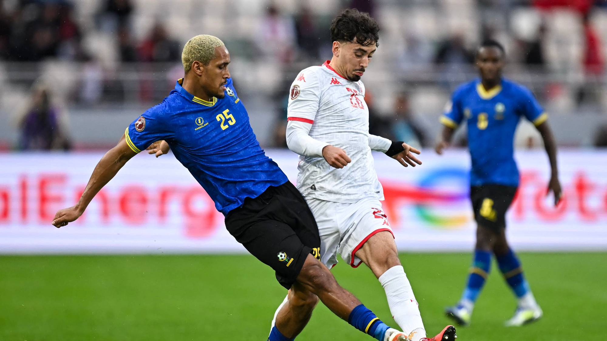 Fotballspillere i aksjon under en kamp. En spiller i blå drakt med nummer 25 prøver å ta ballen fra en spiller i hvit drakt. En tredje spiller i blå drakt sees i bakgrunnen. Stadionet har publikum på tribunen. (Bildebeskrivelsen er laget av en KI-tjeneste)
