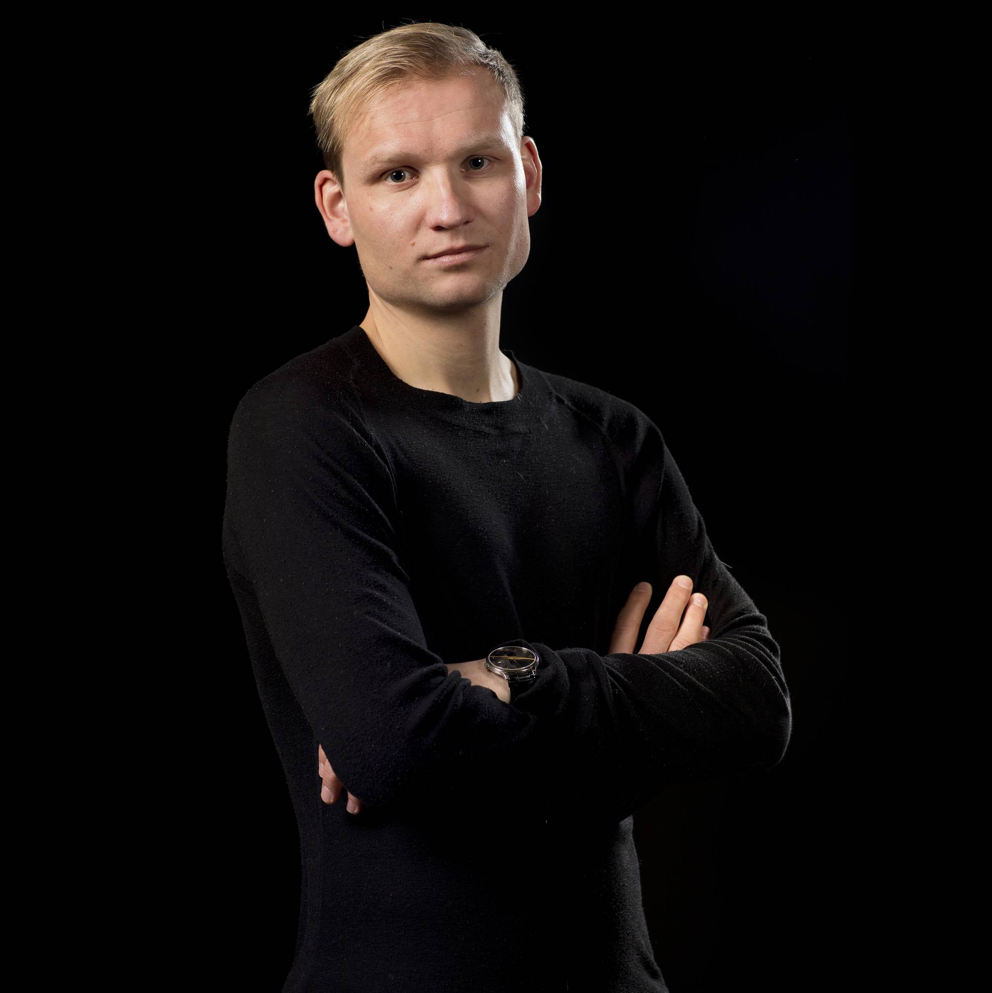 Portrett av Birger Løfaldli. Han har på seg sort genser, som nesten går i ett med den sorte bakgrunnen. Han har lyst, kort hår, har armene i kors og ser alvorlig inn i kameraet.