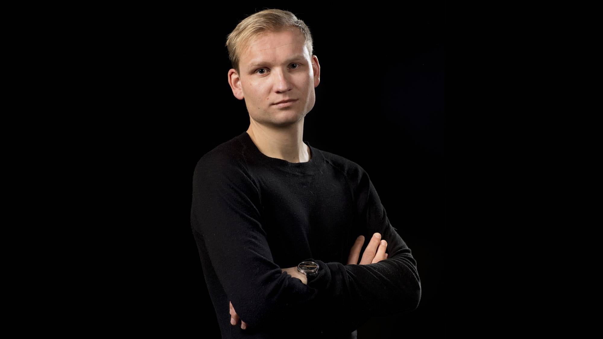 Portrett av Birger Løfaldli. Han har på seg sort genser, som nesten går i ett med den sorte bakgrunnen. Han har lyst, kort hår, har armene i kors og ser alvorlig inn i kameraet.