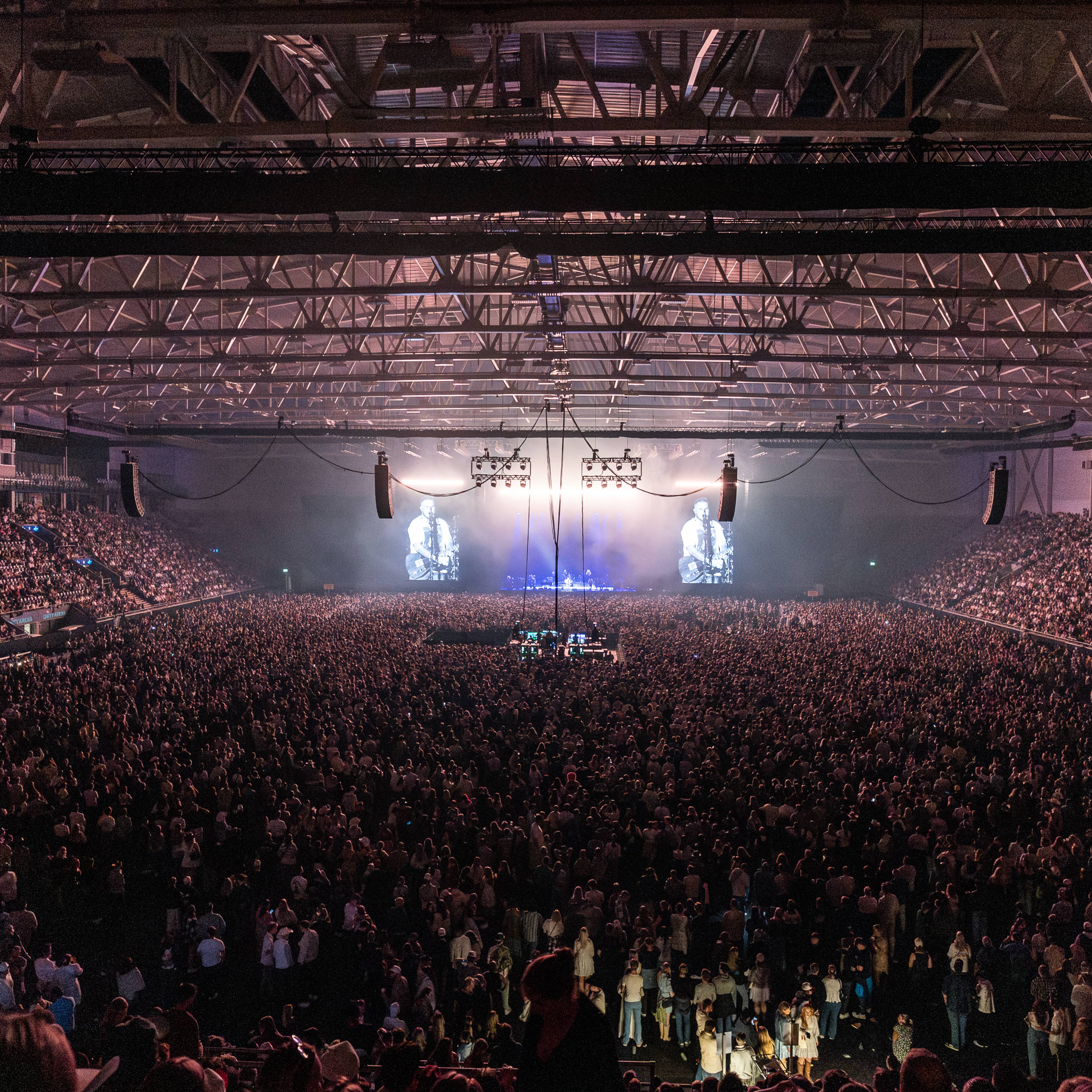 En enorm menneskemengde er samlet på en innendørs arena for å se en konsert. Det er en scene med skjermer som viser bilder av artisten. Publikum fyller både gulvet og tribunene. (Bildebeskrivelsen er laget av en KI-tjeneste)