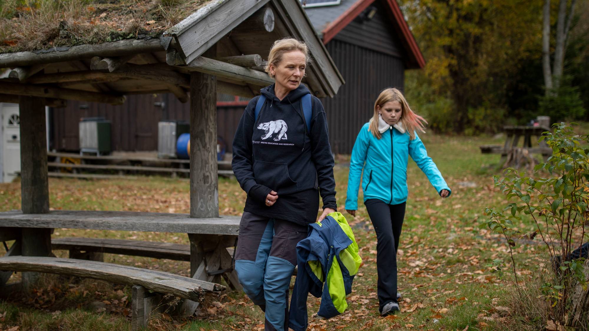 Kvinne og jente går sammen i et skogsterreng foran en trebygning med torvtak. (Bildebeskrivelsen er laget av en KI-tjeneste)