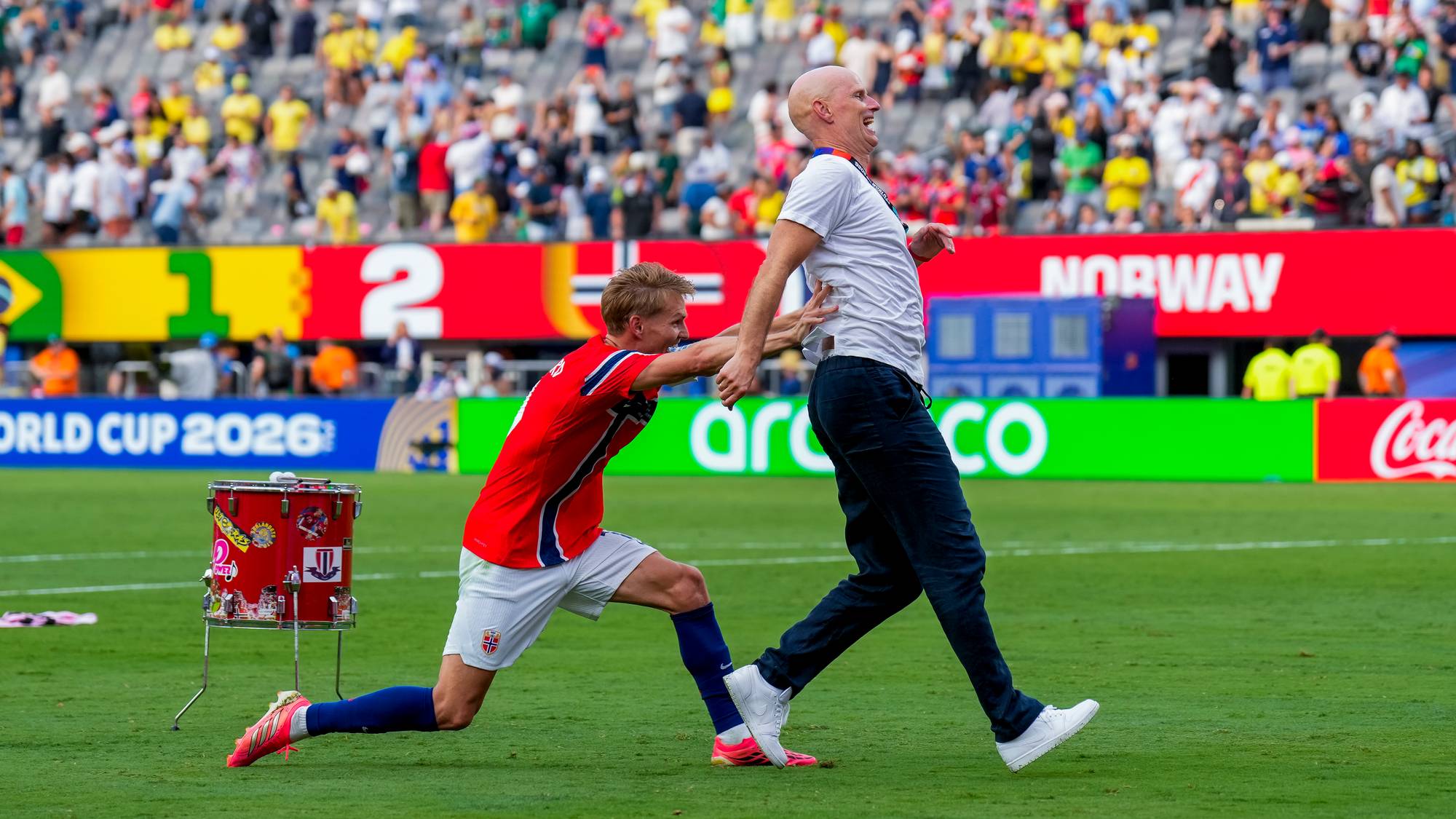 Fotballspiller i norsk landslagsdrakt holder en mann i hvit t-skjorte på en fotballbane. (Bildebeskrivelsen er laget av en KI-tjeneste)