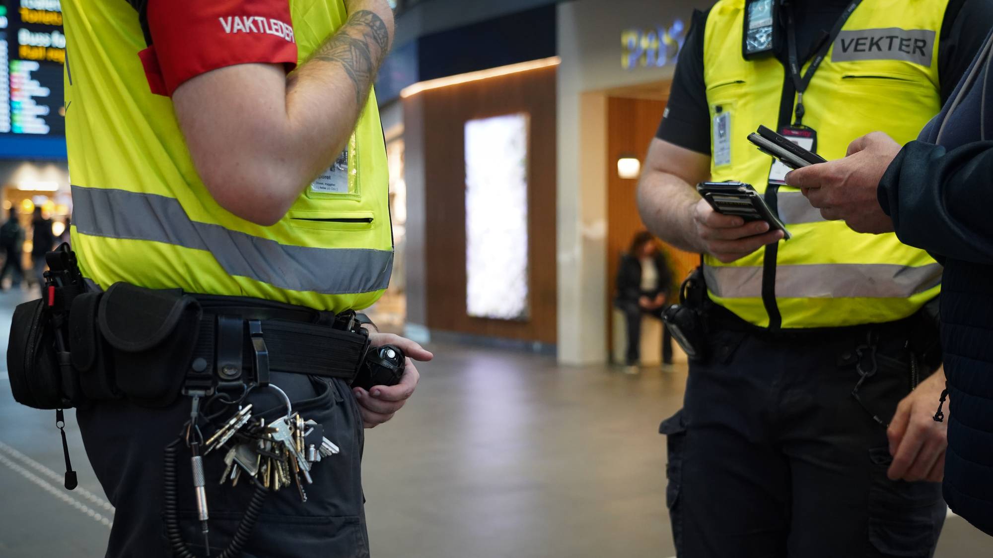 To vektere i gule vester med refleksstriper ser på telefoner. En av dem har en nøkkelknippe hengende fra beltet. I bakgrunnen sitter en person. (Bildebeskrivelsen er laget av en KI-tjeneste)