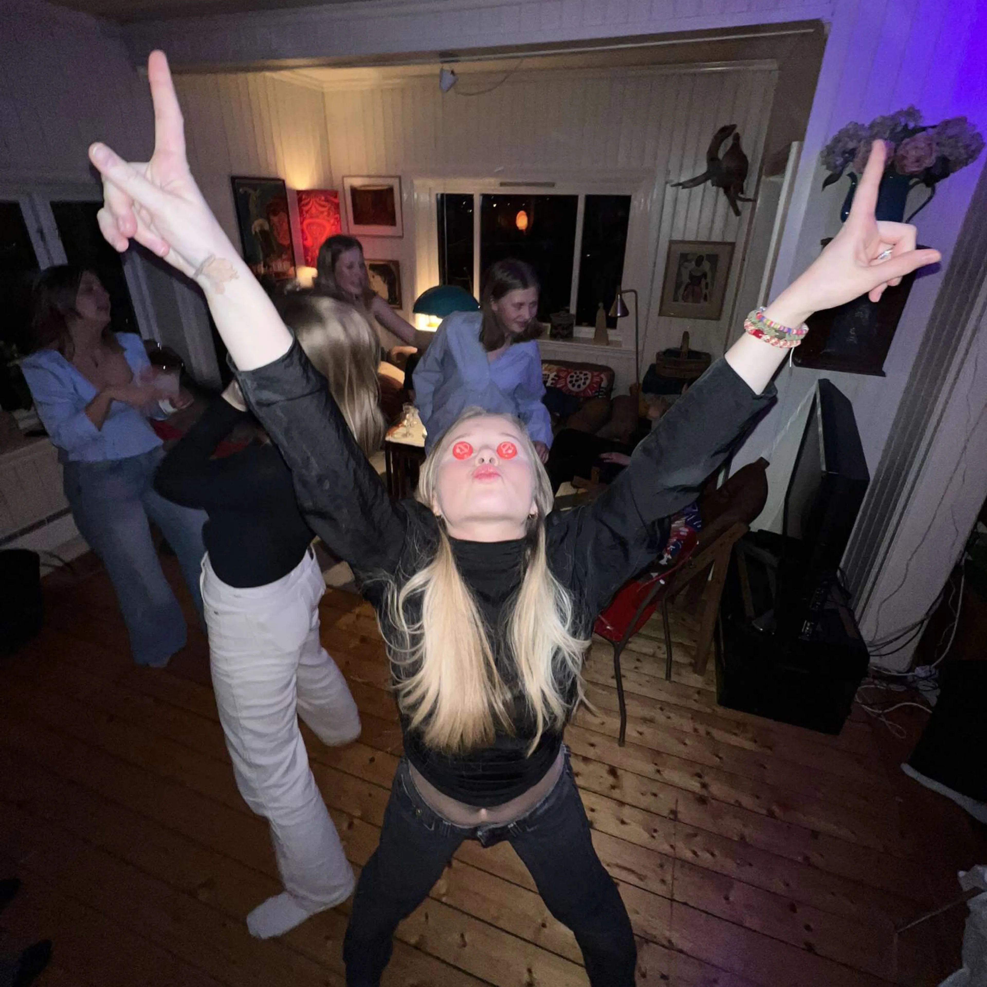 En kvinne med en rød flaskekork på hvert øye står i en stue med venner som danser i bakgrunnen. (Bildebeskrivelsen er laget av en KI-tjeneste)