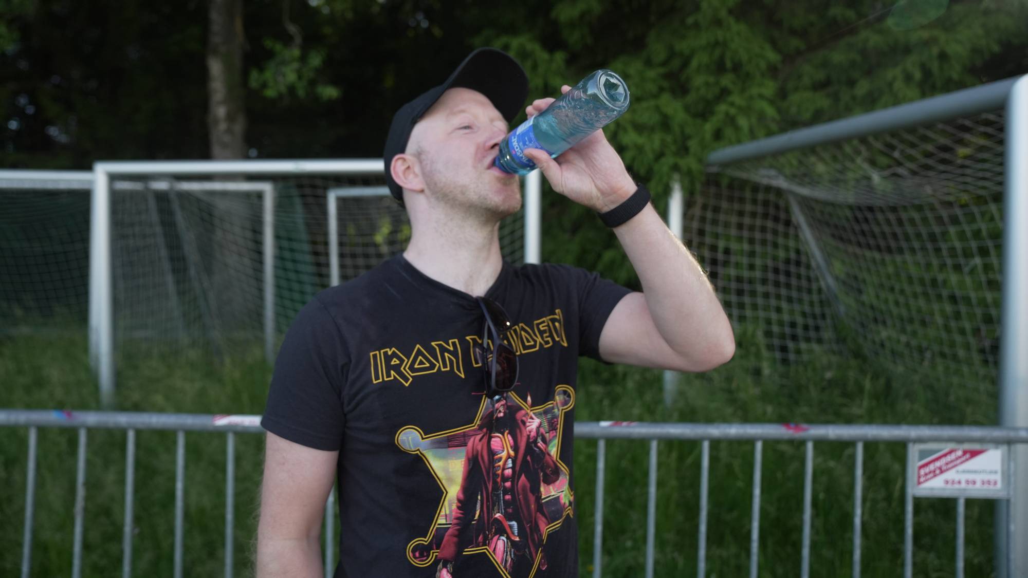 Mann i Iron Maiden-t-skjorte drikker vann fra en flaske foran et fotballmål. 