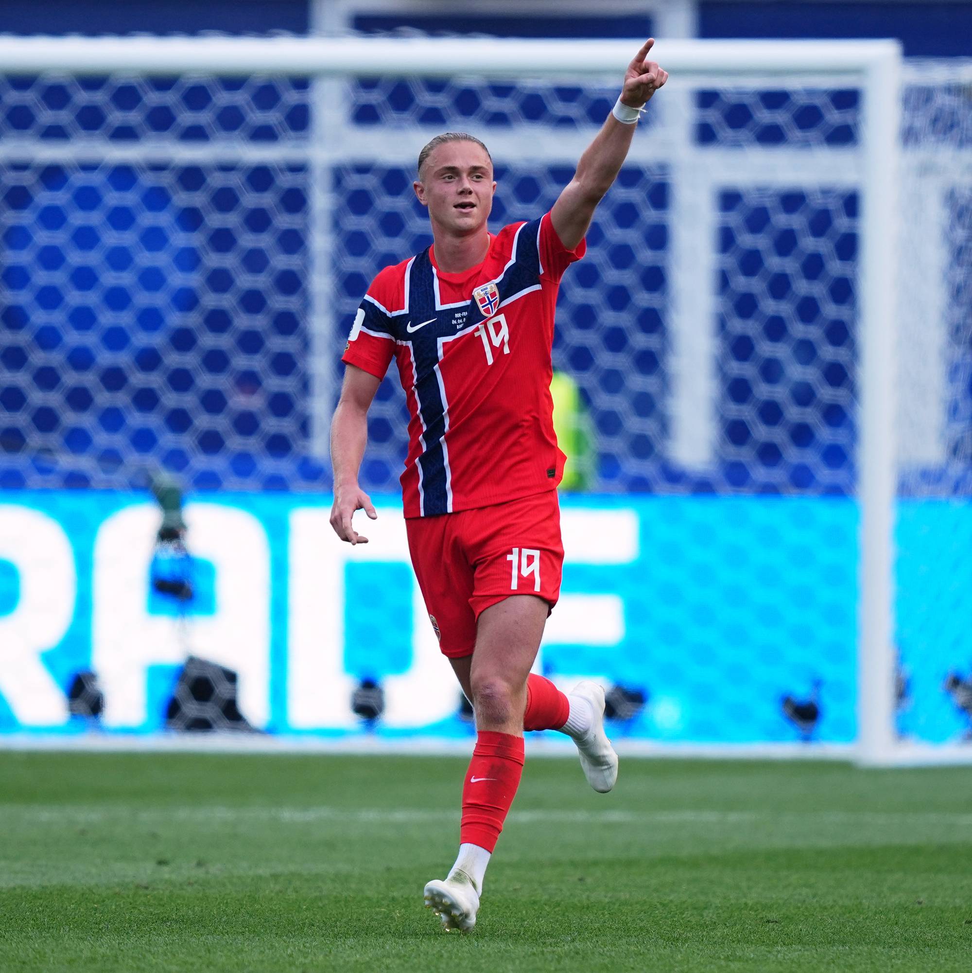 Norsk fotballspiller i rød landslagsdrakt jubler med fingeren i været foran et fotballmål. (Bildebeskrivelsen er laget av en KI-tjeneste)
