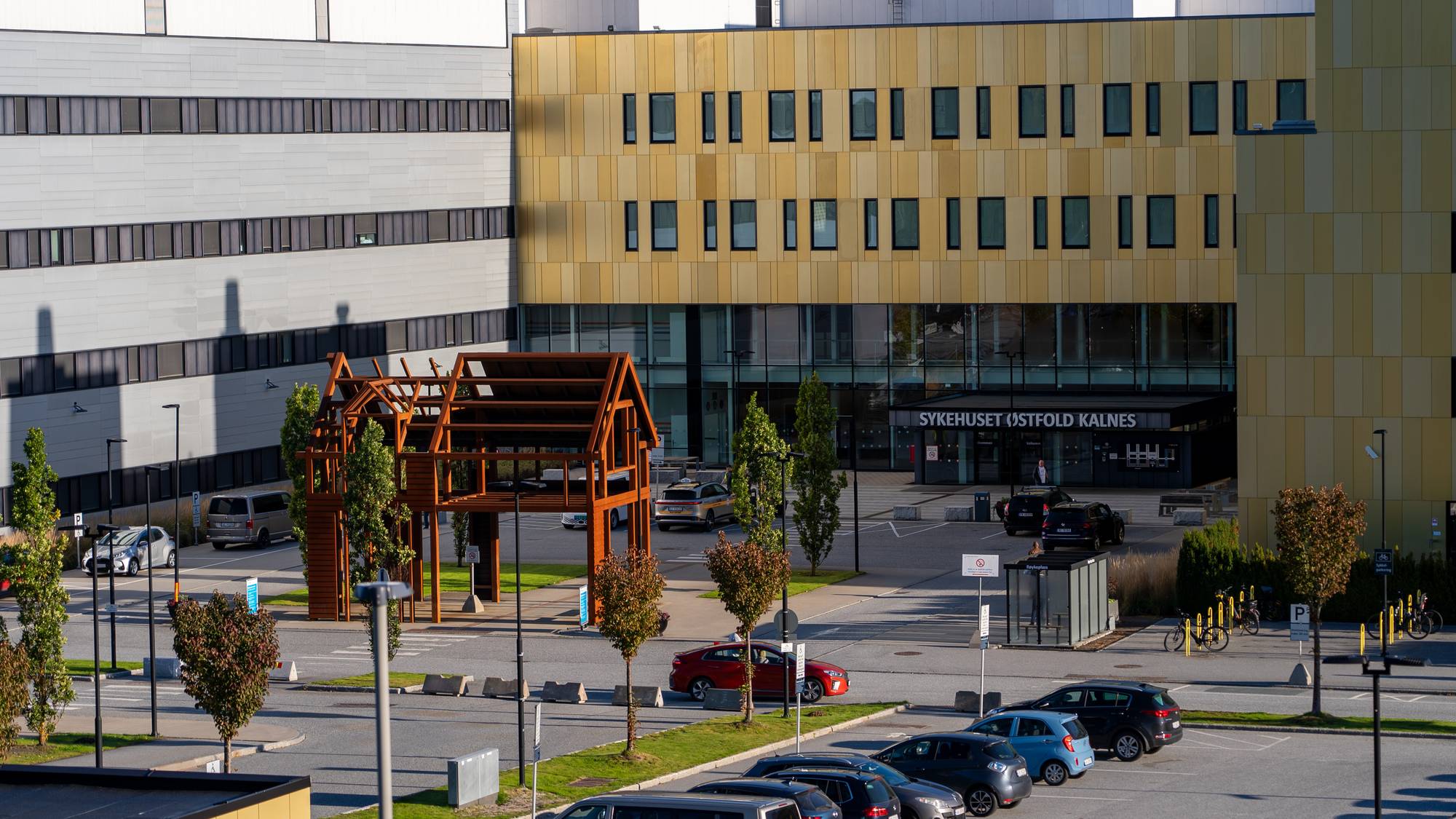 Sykehuset Østfold Kalnes har en gul fasade og store vindusflater. Utenfor sykehuset er det en parkeringsplass med flere biler. Det er også et utendørs trebygg. (Bildebeskrivelsen er laget av en KI-tjeneste)