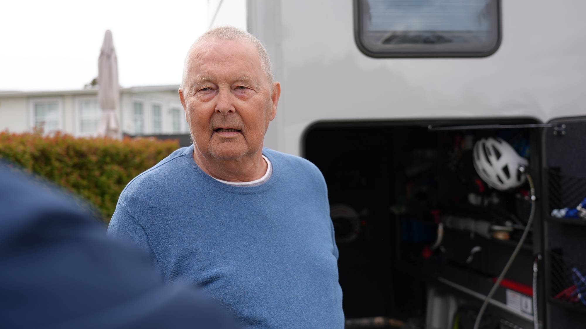 Aage Pedersen har en blå genser og ser til venstre for kamera. Bak han er bobilen med sykler i. 