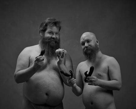 To menn med skjegg står nakne og holder opp sexleketøy. Han ene er kraftig bygd med stor mage. Han andre er skallet. Begge smiler.