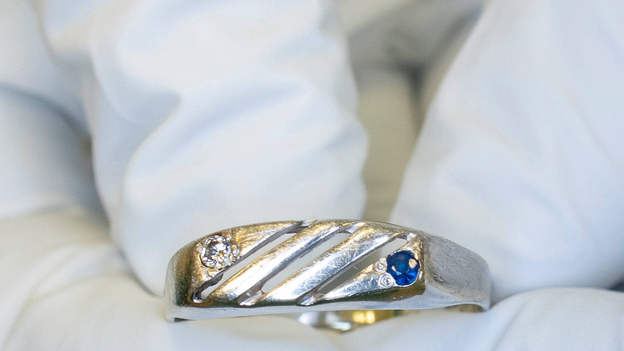 Ring med blå og hvit sten, sølvfarget. Holdes av hånd med hvit hanske 