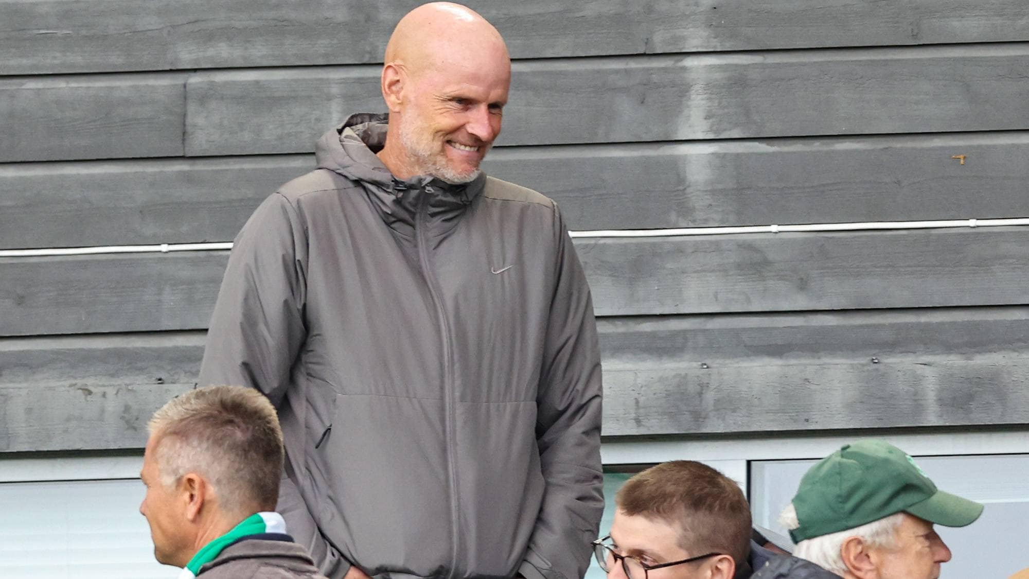 Kjetil Rekdal smiler mens han står på tribunen omgitt av andre tilskuere. (Bildebeskrivelsen er laget av en KI-tjeneste)
