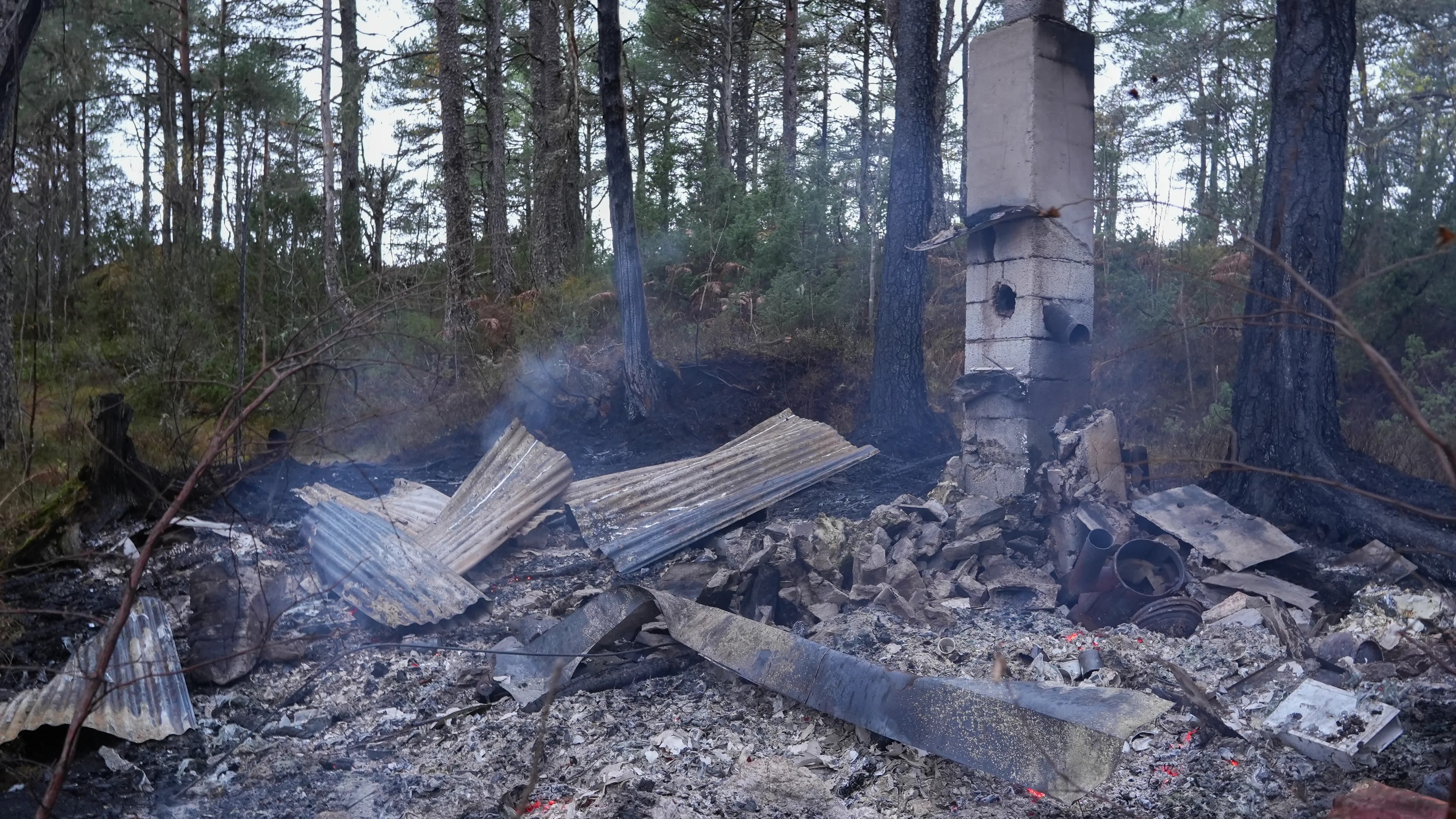 Brannruin etter et nedbrent bygg i skogsterreng. En frittstående pipe står igjen blant aske og metall. (Bildebeskrivelsen er laget av en KI-tjeneste)