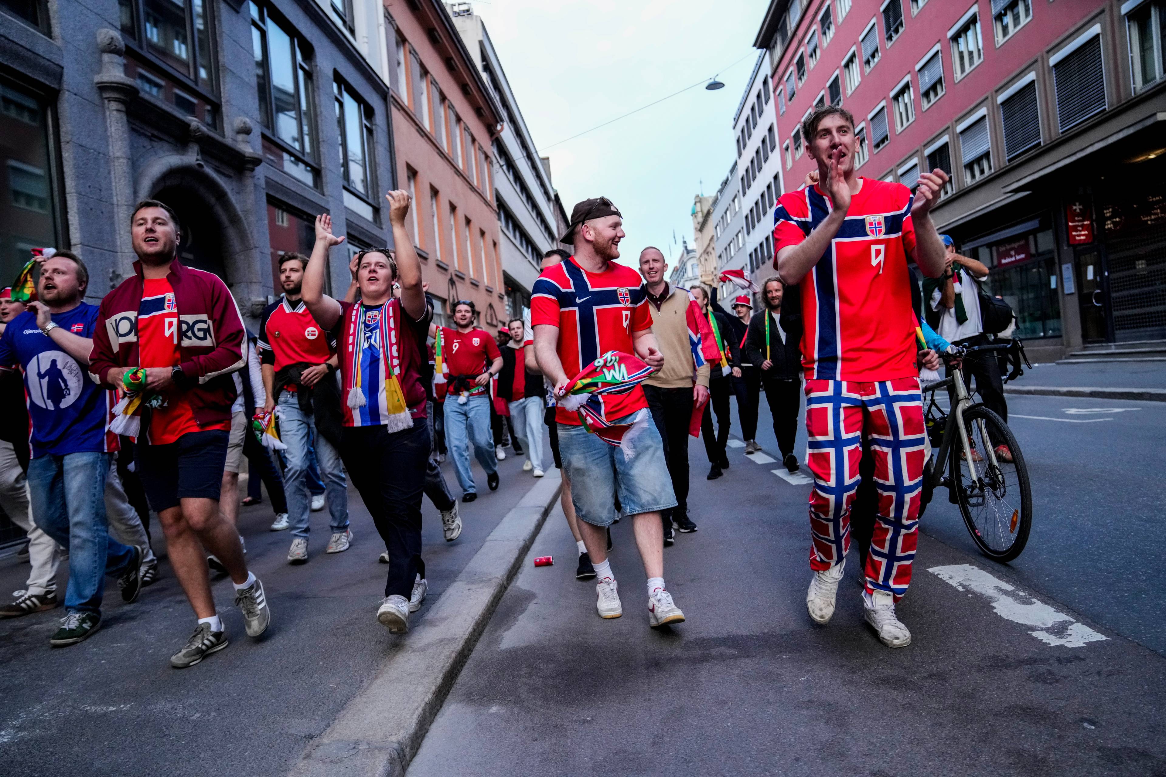 En gruppe menn i norske fotballdrakter går i en gate. Noen vifter med armene og roper. En sykkel står parkert ved siden av veien. Gaten er omkranset av bygninger.