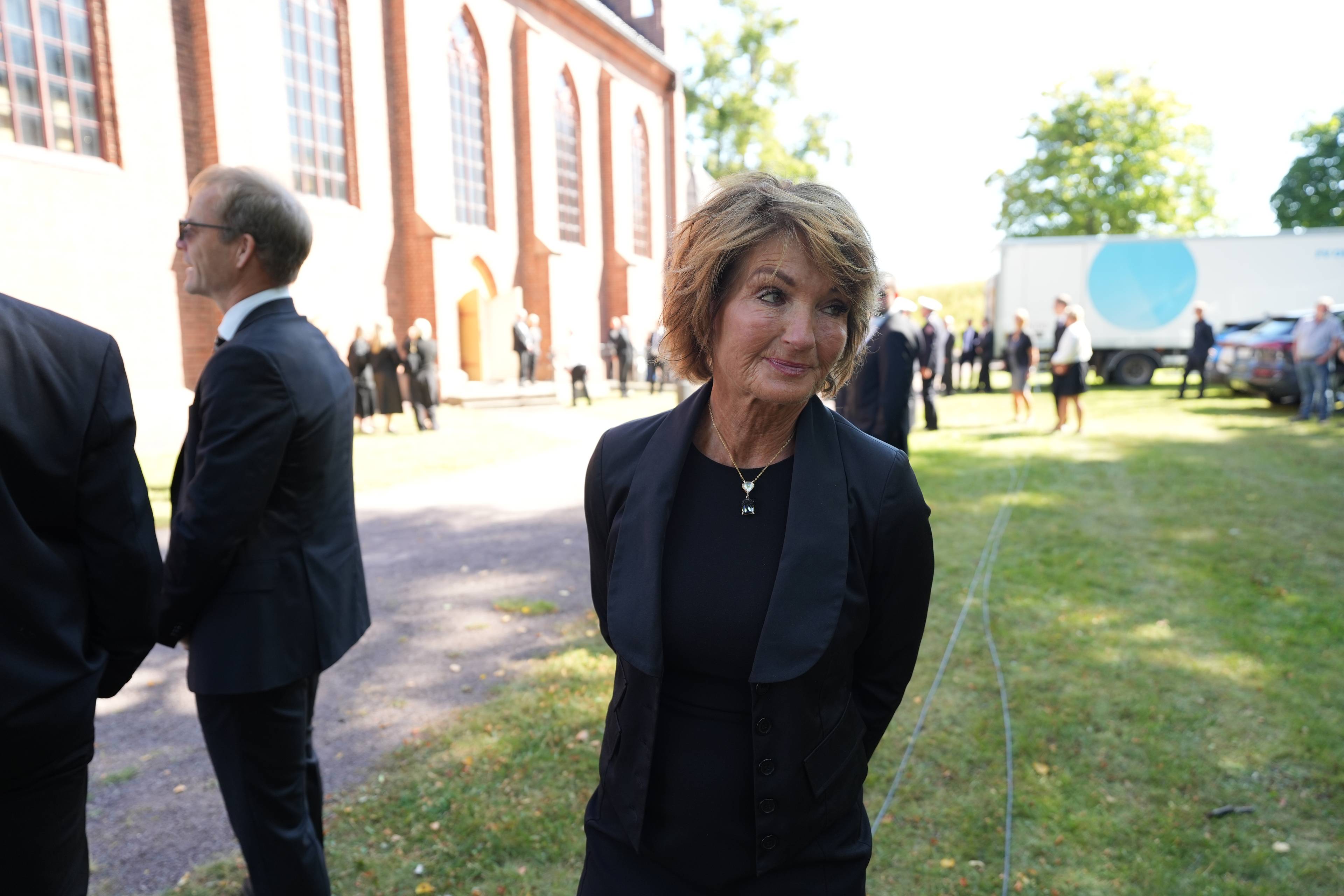 Kvinne i svart kåpe foran en kirke. Flere personer i dress og svarte klær står i bakgrunnen.