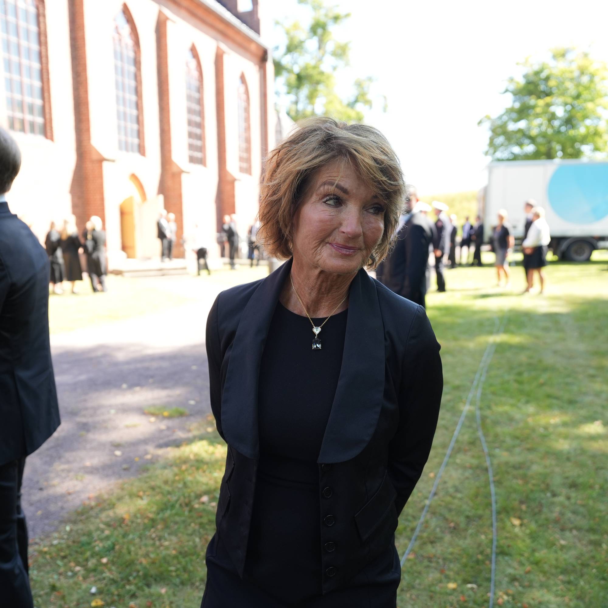 Kvinne i svart kåpe foran en kirke. Flere personer i dress og svarte klær står i bakgrunnen.