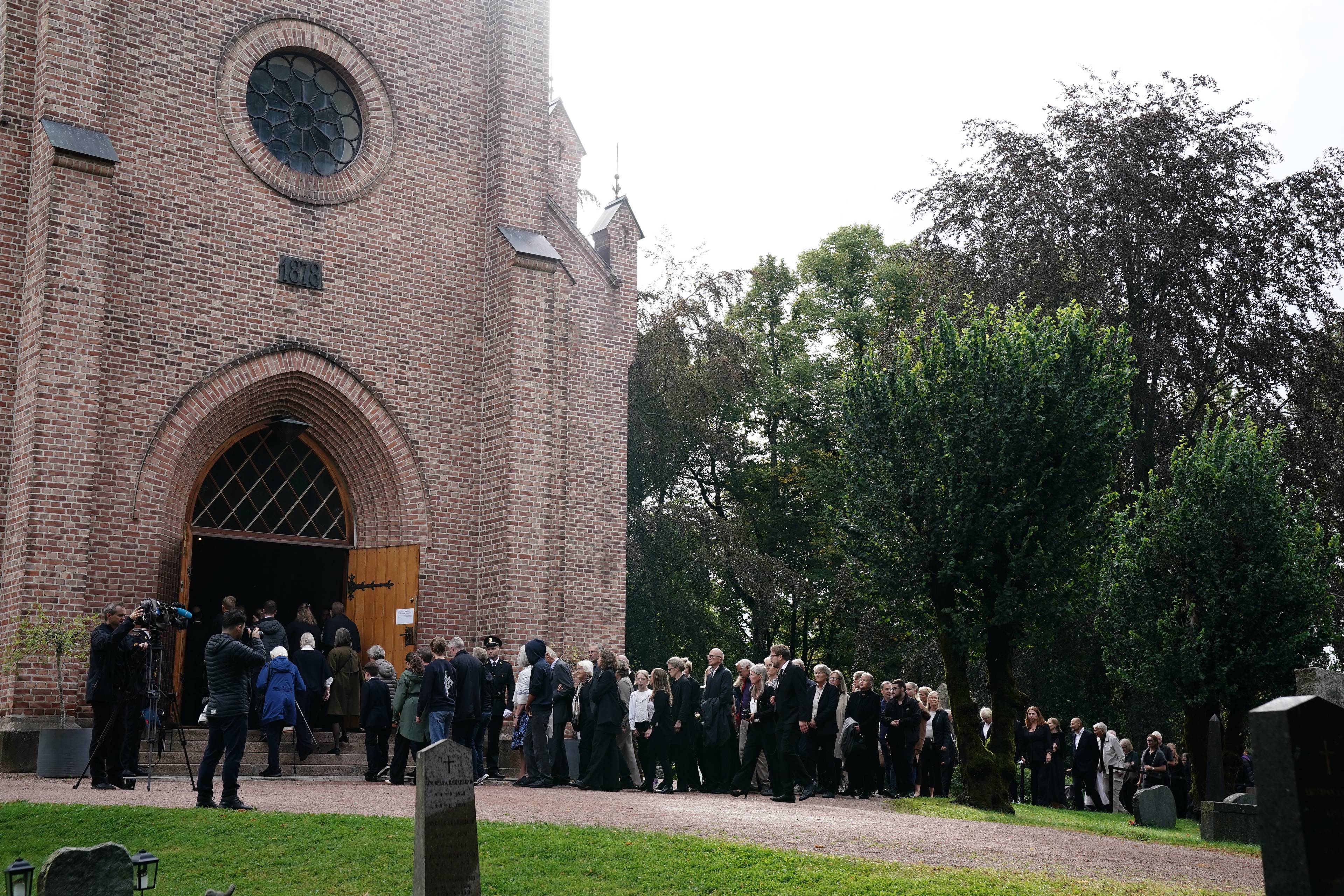 Folk i mørke klær står i kø utenfor en kirke før en sørgegudstjeneste.