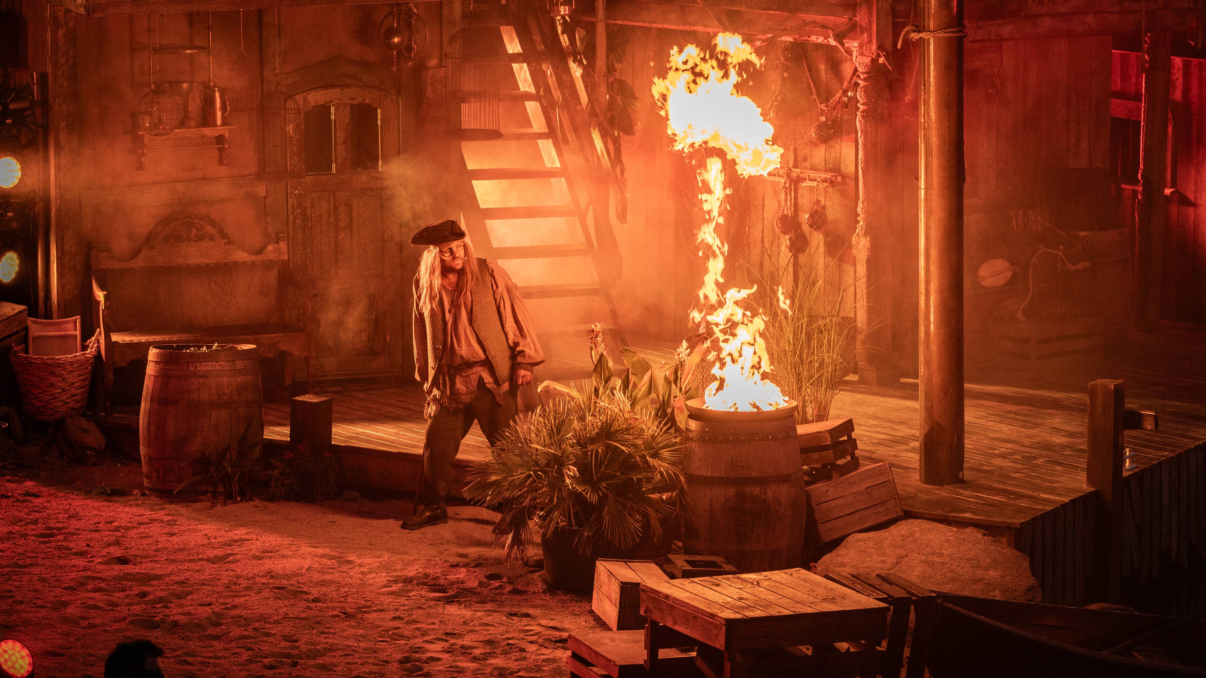 Skuespiller i piratkostyme på teaterscene med flammer fra en tønne i rødt lys. (Bildebeskrivelsen er laget av en KI-tjeneste)
