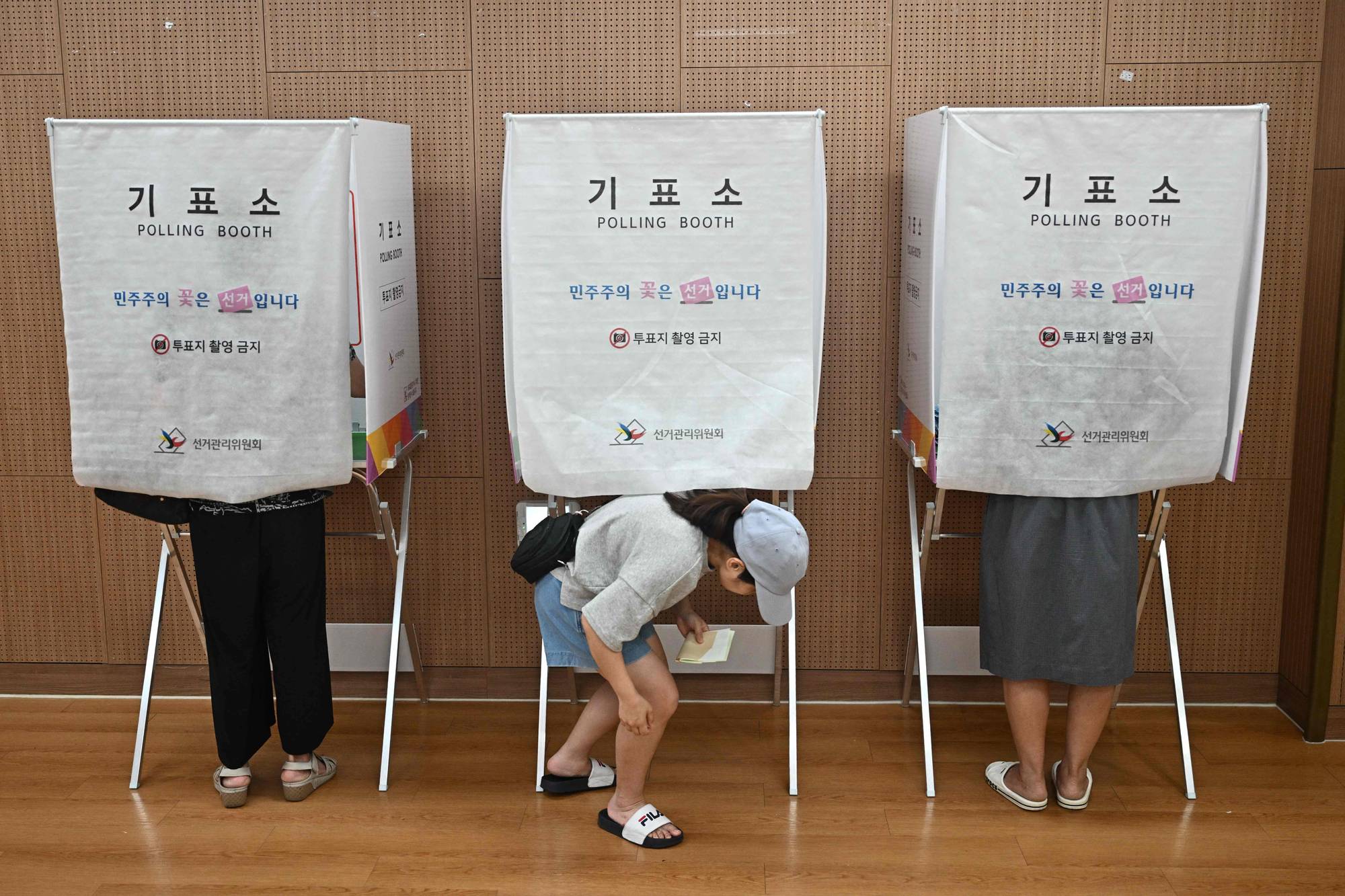 Folk stemmer i valgbokser. En person bøyer seg ned for å fylle ut stemmeseddel. Bildet viser tre valgbokser med skilt på koreansk og engelsk.