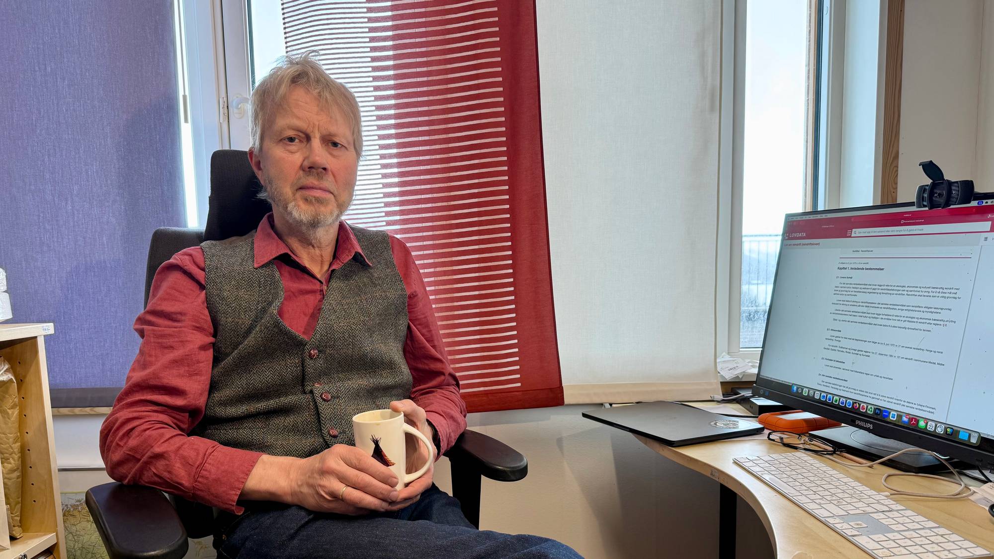 En mann i rød skjorte og brun vest holder et kaffekrus i hånda