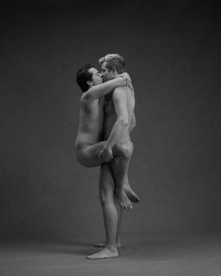 En naken mann står på gulvet og løfter en annen naken mann mens de kysser