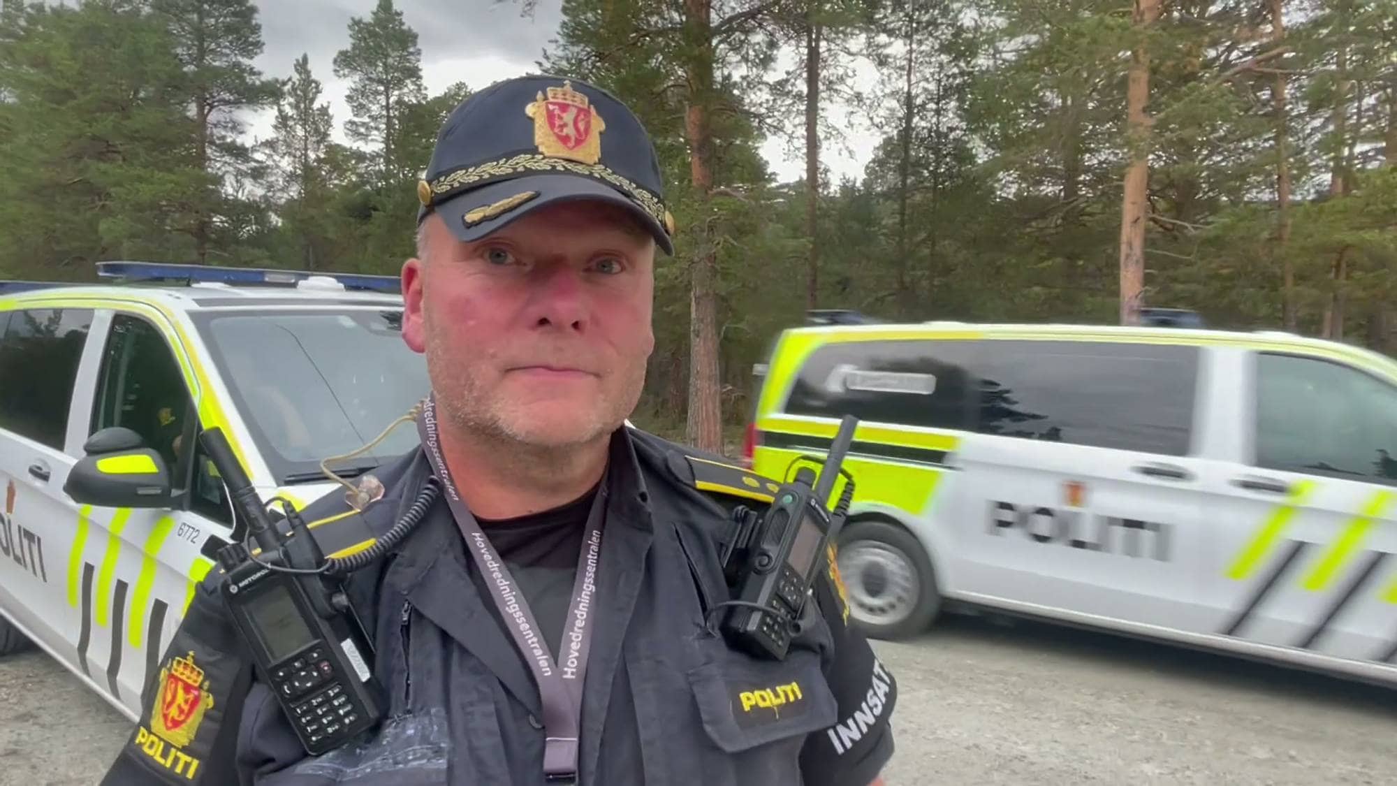 Politi i uniform foran to politibiler i et skogsområde. (Bildebeskrivelsen er laget av en KI-tjeneste)