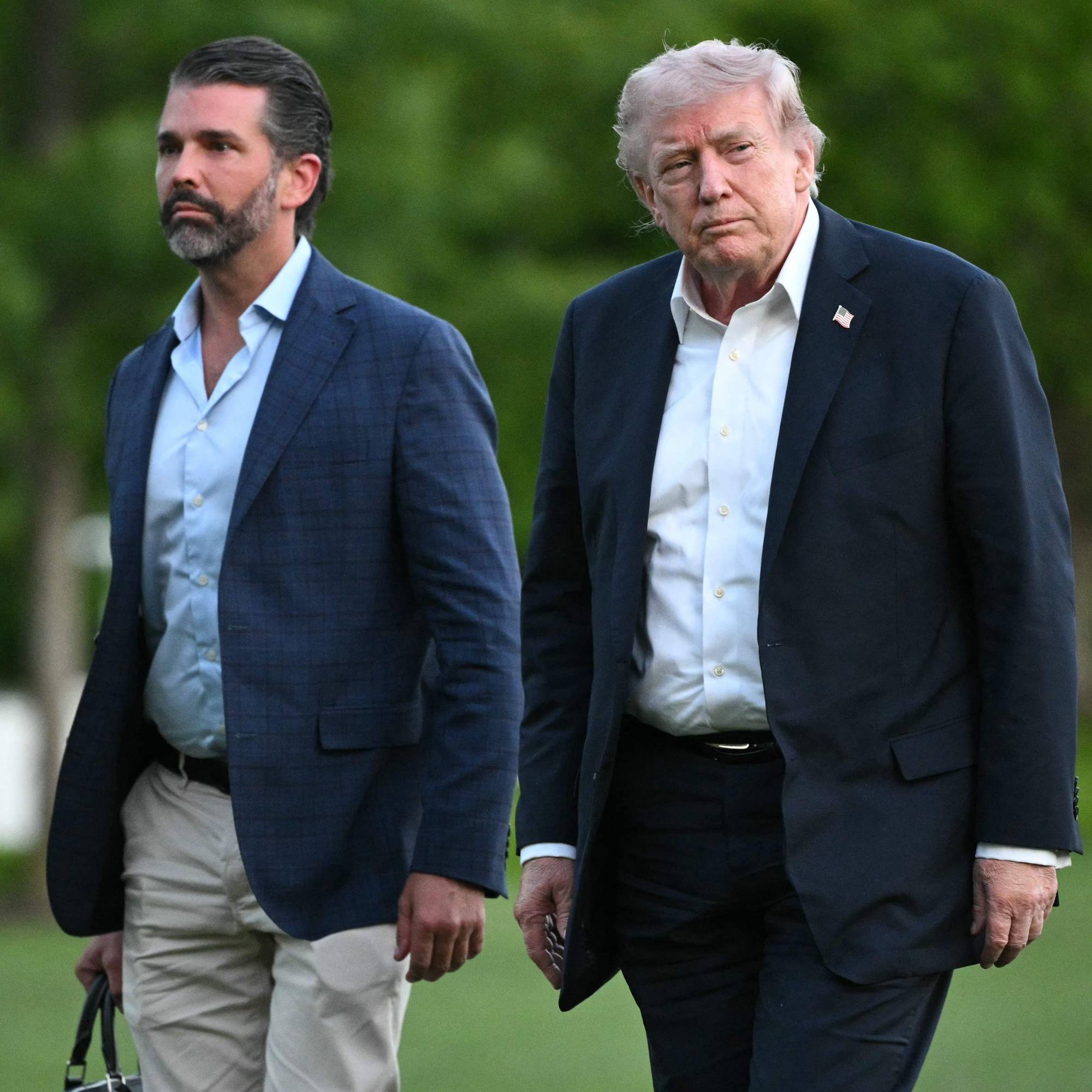 Donald Trump og Donald Trump Jr. går på et grønt område. Begge mennene er kledd i jakker og skjorter. Trump Jr. bærer en bag i venstre hånd. (Bildebeskrivelsen er laget av en KI-tjeneste)