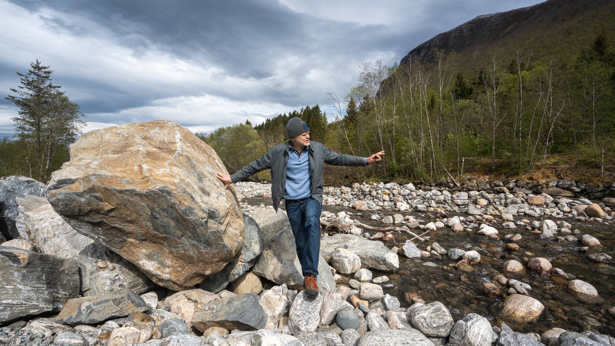 En mann med grå lue, blå genser og jeans står på steiner ved en elv. Han har en arm mot en stor stein og peker med den andre mot elva. Skyet vær med mørke skyer dominerer himmelen. (Bildebeskrivelsen er laget av en KI-tjeneste)